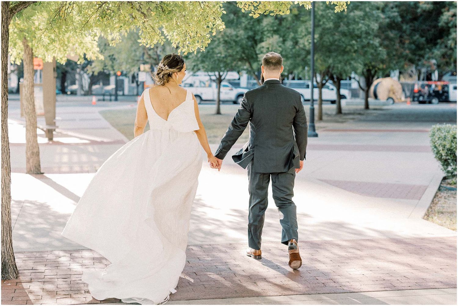 Wedding couple walking together on a sunlit sidewalk with trees in background.