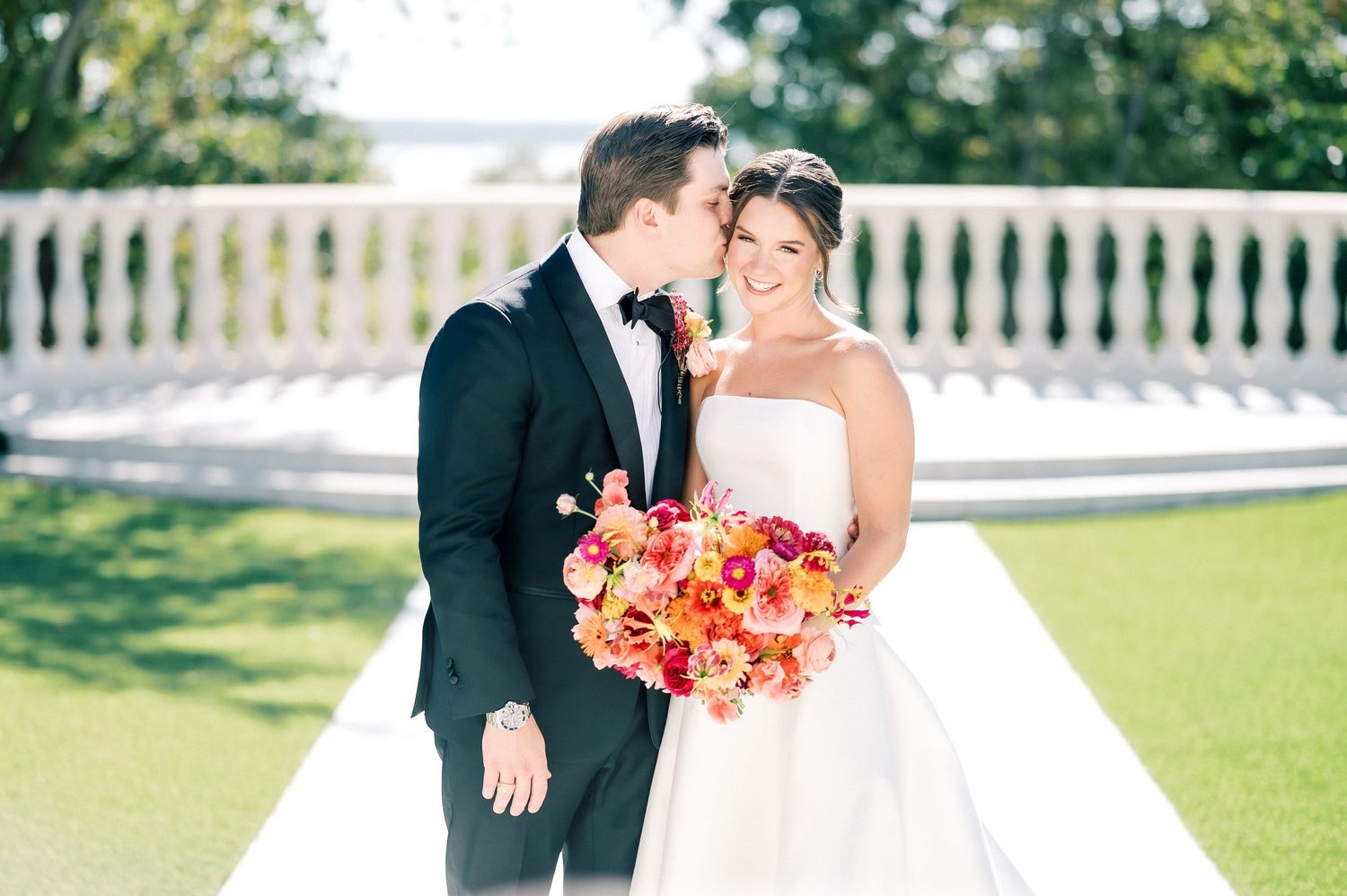 Wedding couple posing with vibrant coral bouquet against white picket fence.