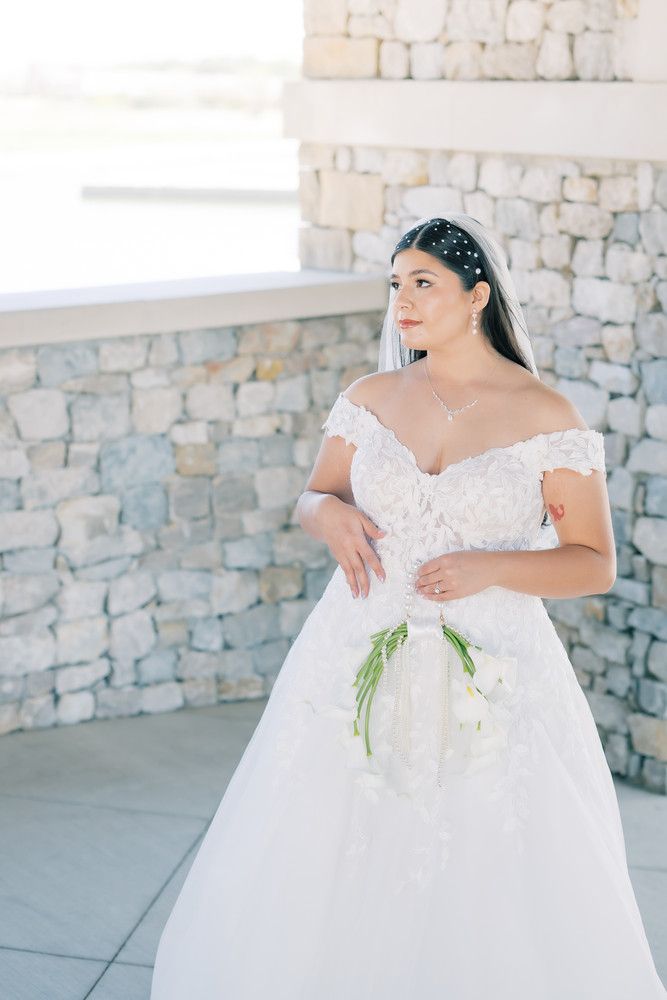 A bride in a classic white ball gown stands against a stone wall holding white flowers.