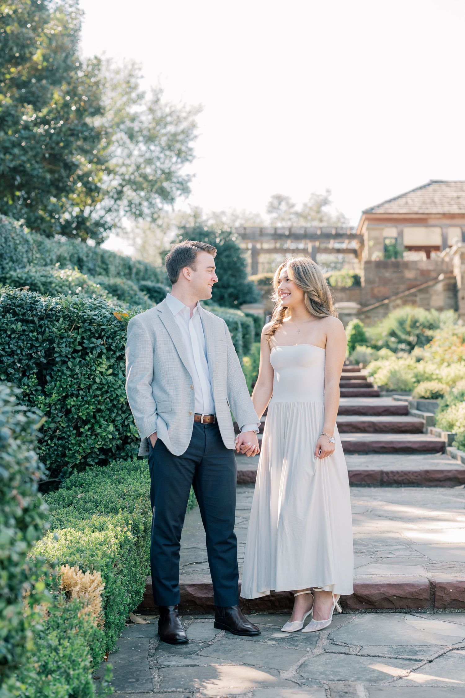 A couple shares a romantic moment together in a garden setting with stone steps and lush greenery in the background.