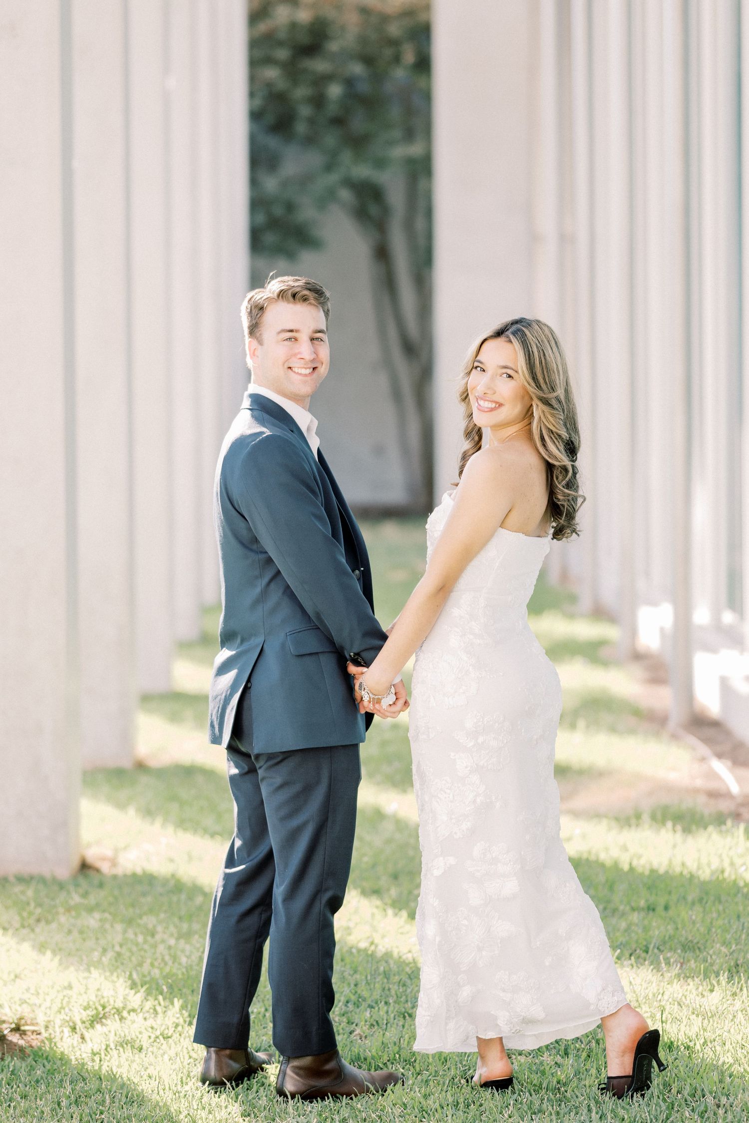 A couple in wedding attire poses between white columns outdoors on a sunny day.