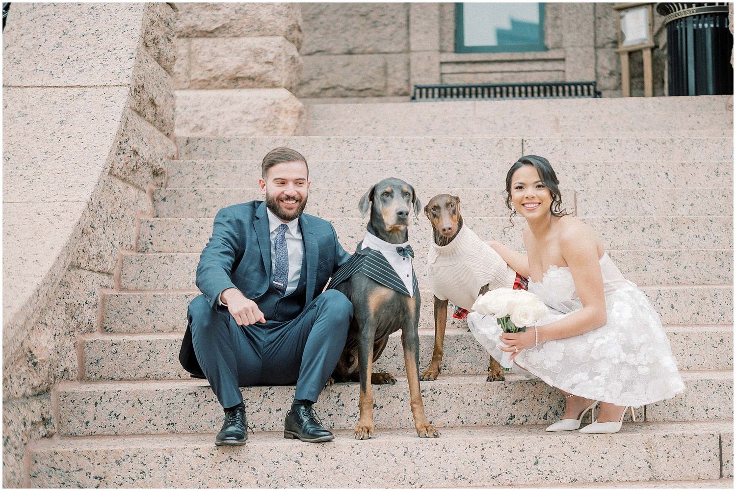 Couple in wedding attire sits with their dog on stone steps outside a building.