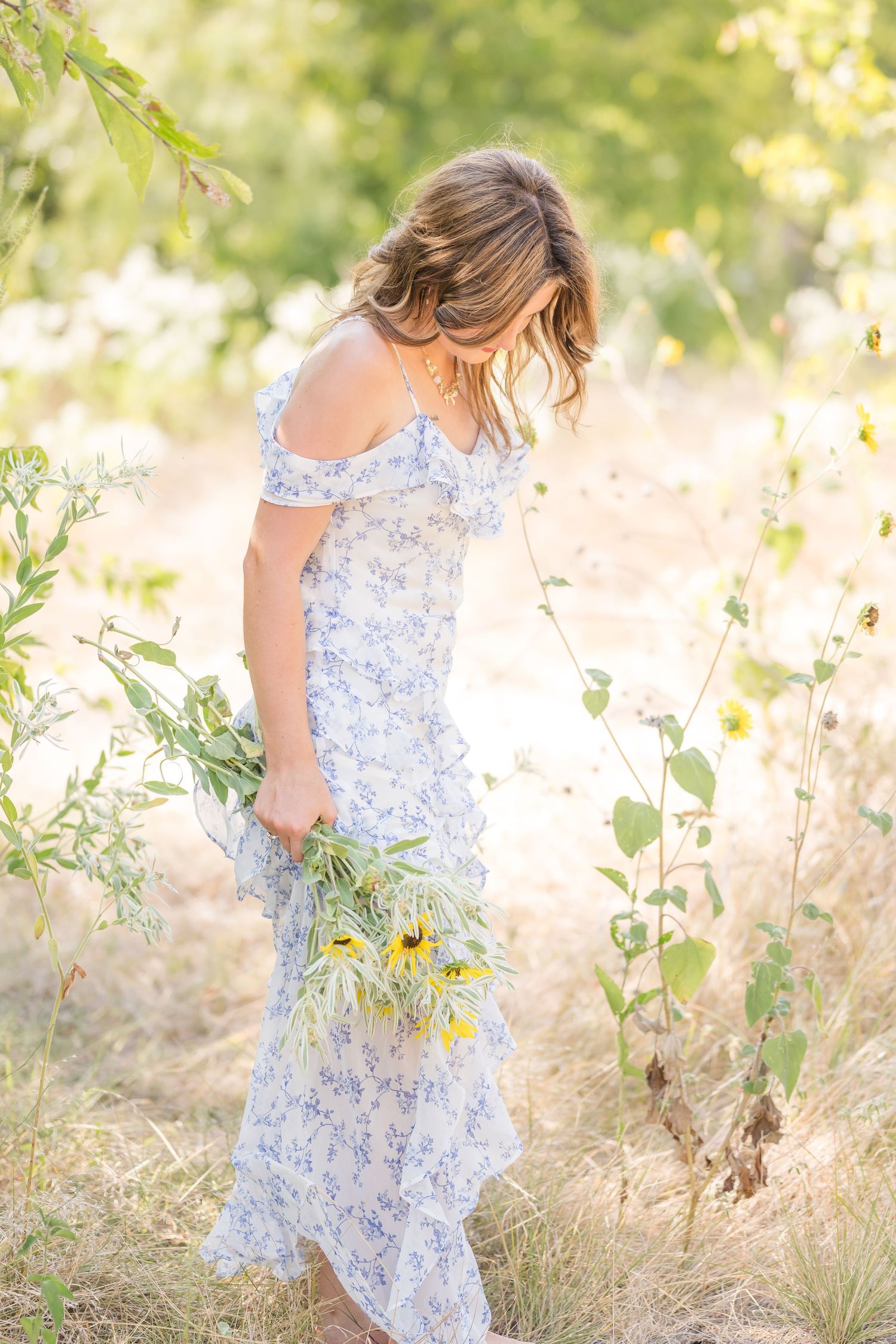 Woman in floral off-shoulder maxi dress holding wildflowers in a sunlit meadow.