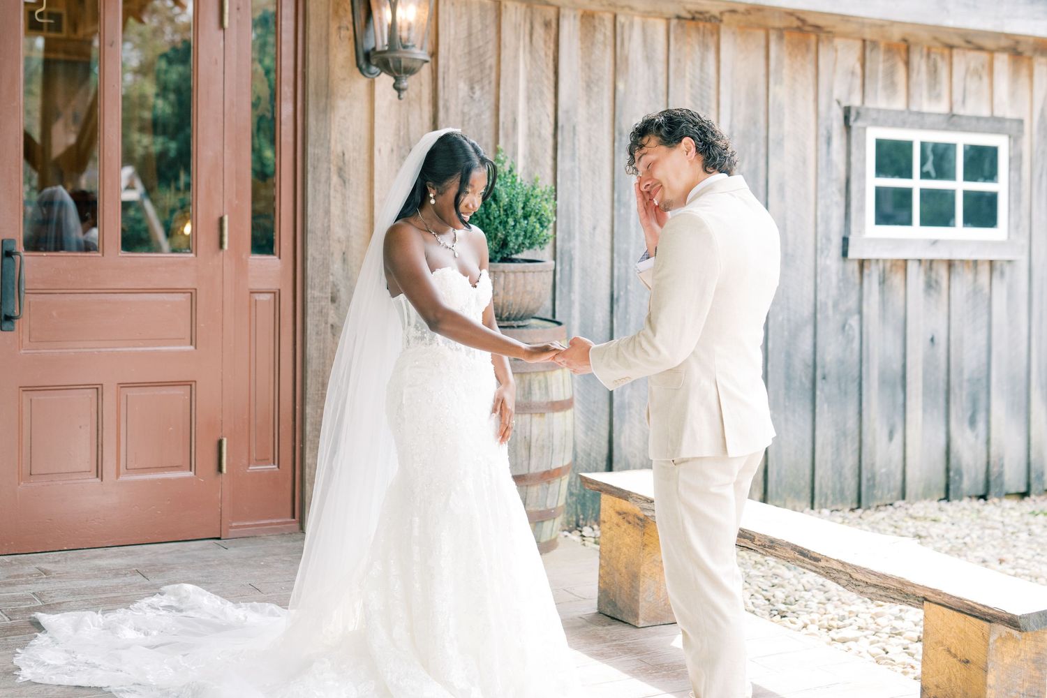 A bride in a white gown and groom in ivory suit share a first look moment outside a rustic barn wedding venue.
