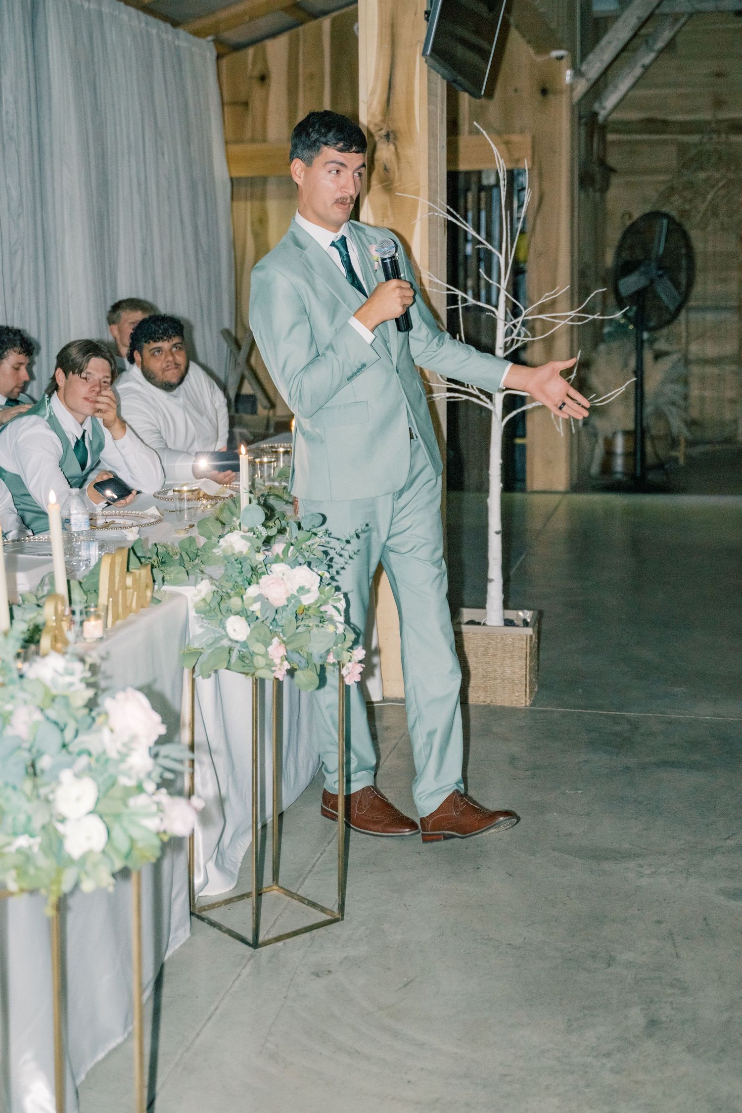 Groomsman in sage green suit delivers speech with microphone at elegant barn wedding reception.