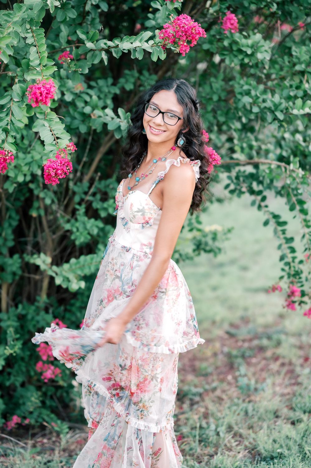 Smiling young woman in floral dress and glasses posing under pink flowering arch in lush green garden.