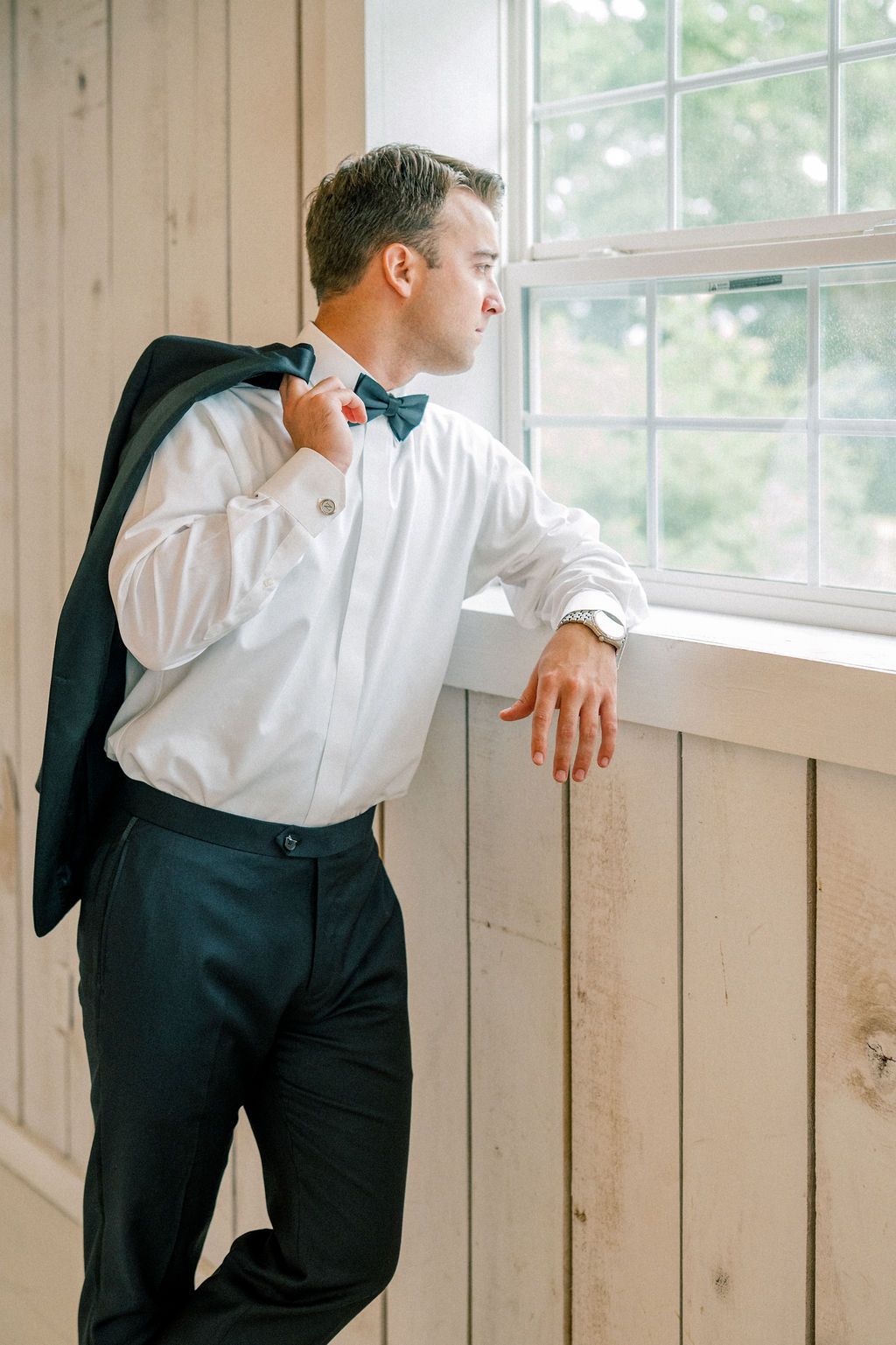 Dapper groom in black tuxedo with teal bow tie holds jacket over shoulder while gazing out a bright window.