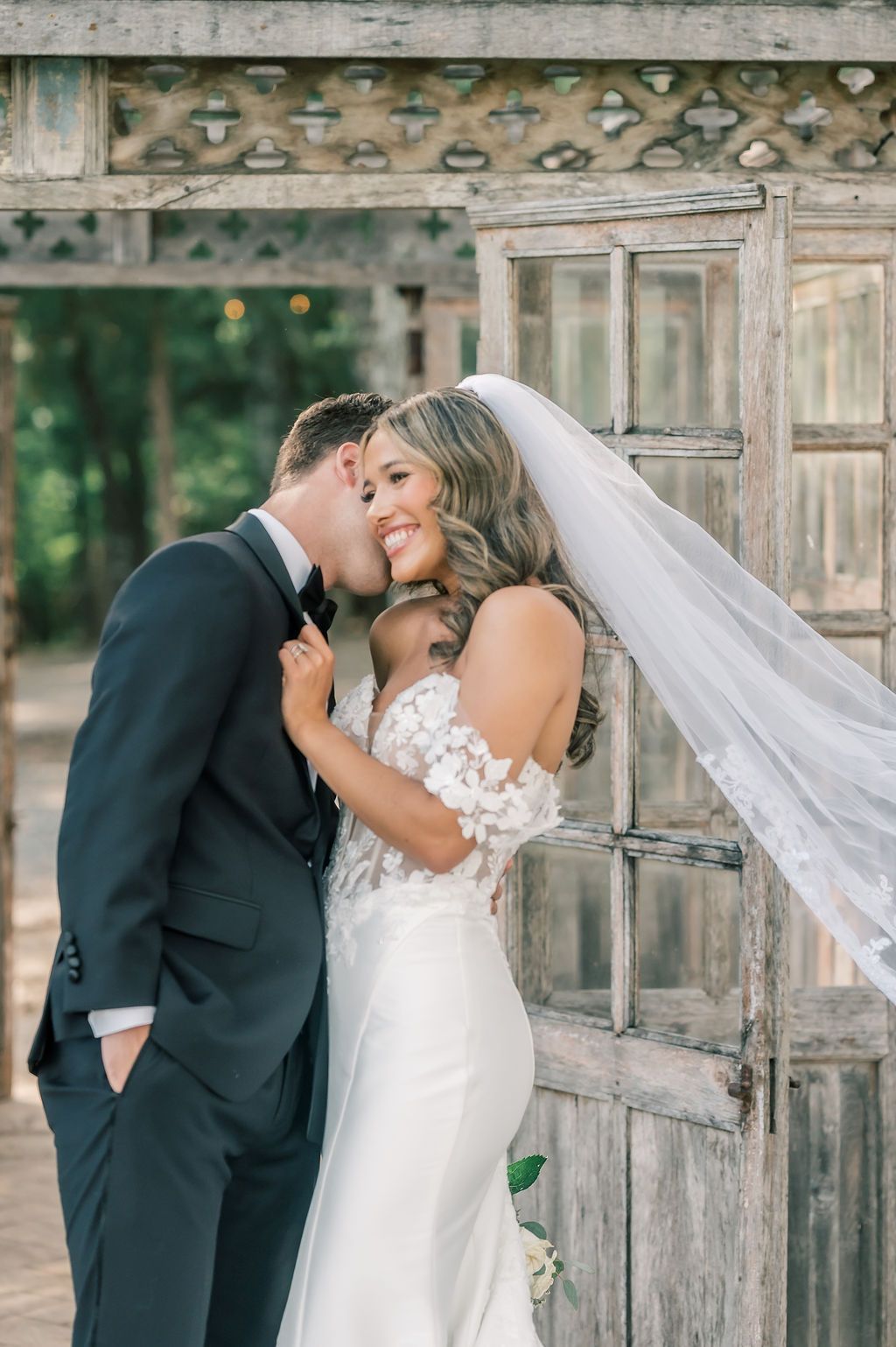Groom in black tux kisses bride in lace gown and veil beside rustic wooden doors at elegant outdoor wedding.
