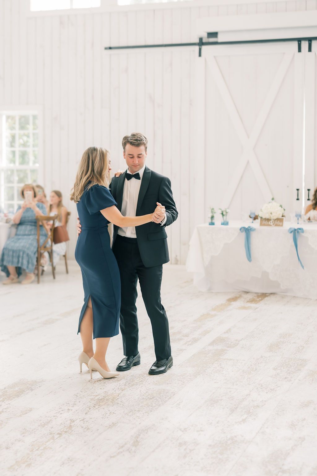 Groom dances with his mother in a navy dress in a bright white barn wedding venue during the reception.