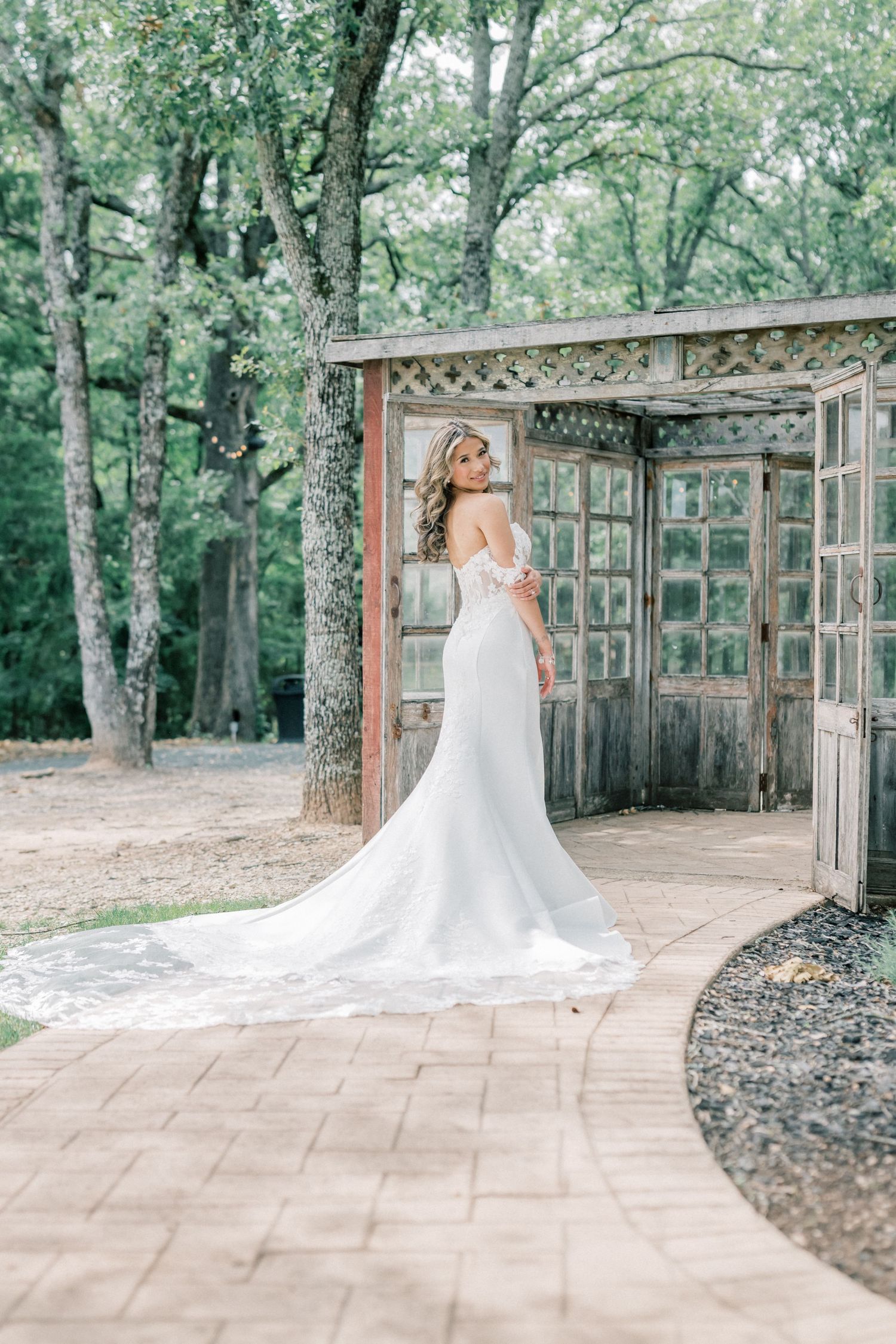 Bride in off-shoulder mermaid gown with train poses at rustic wooden door arch outdoors.