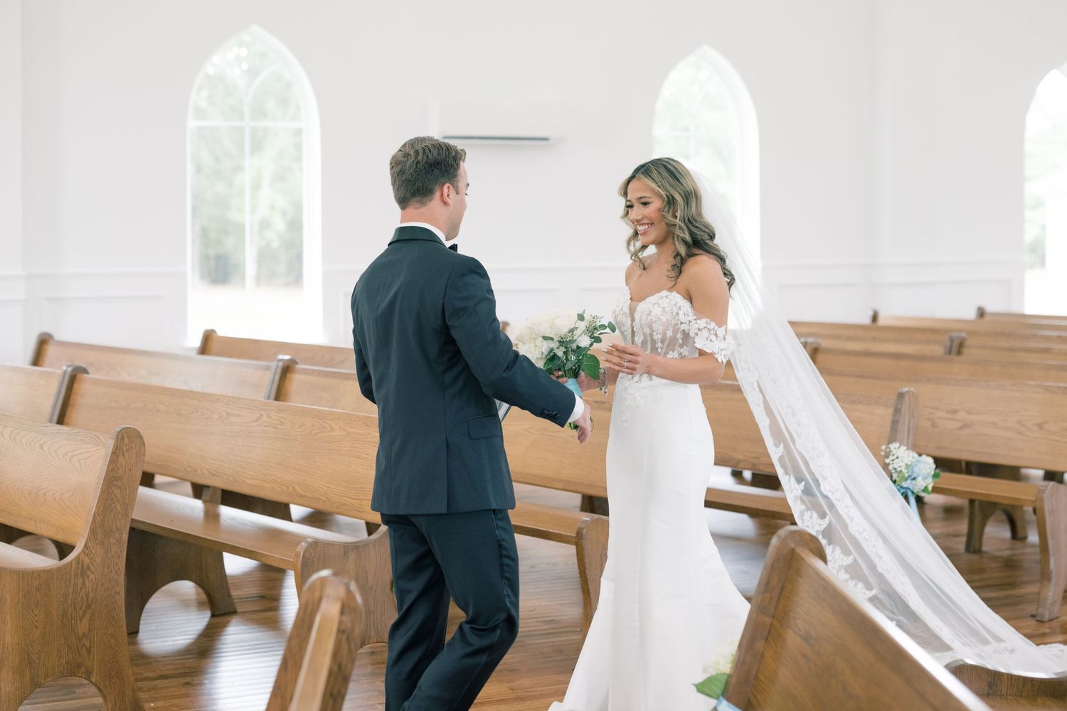 Bride and groom share a tender first look moment inside a bright white chapel with wooden pews.