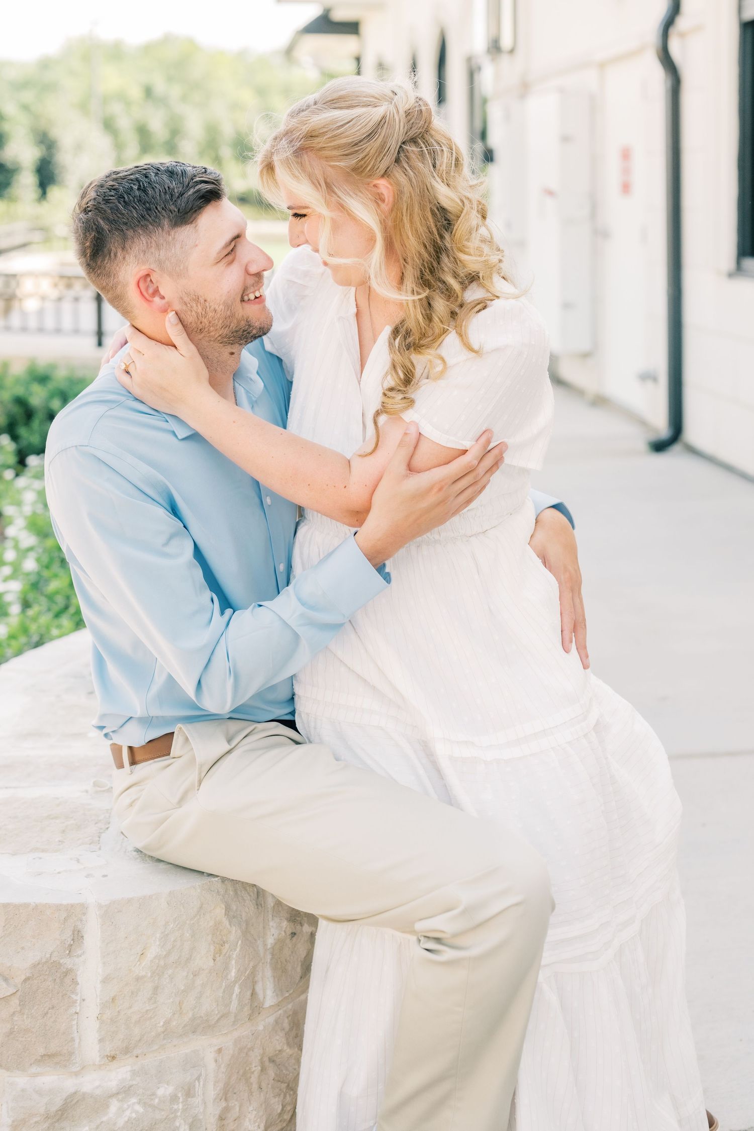 A smiling couple embraces outdoors, woman in white dress sits on stone wall with man in light blue shirt.