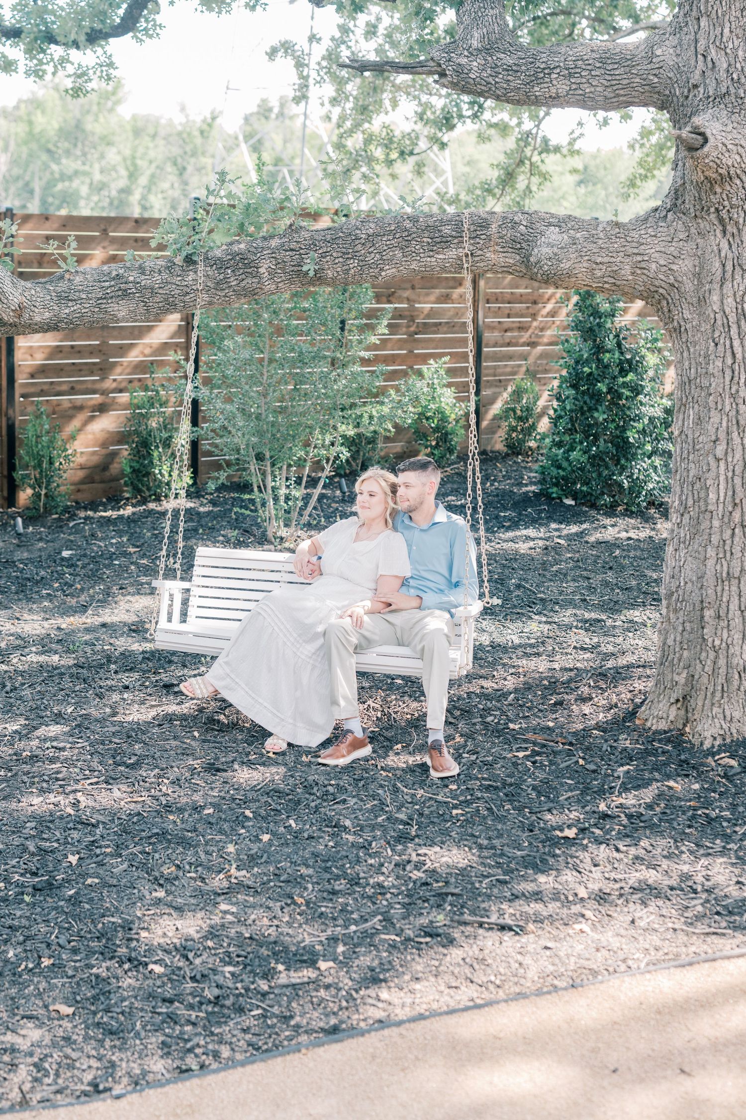 Couple sitting together on a white wooden swing hanging from a large oak tree in a garden.