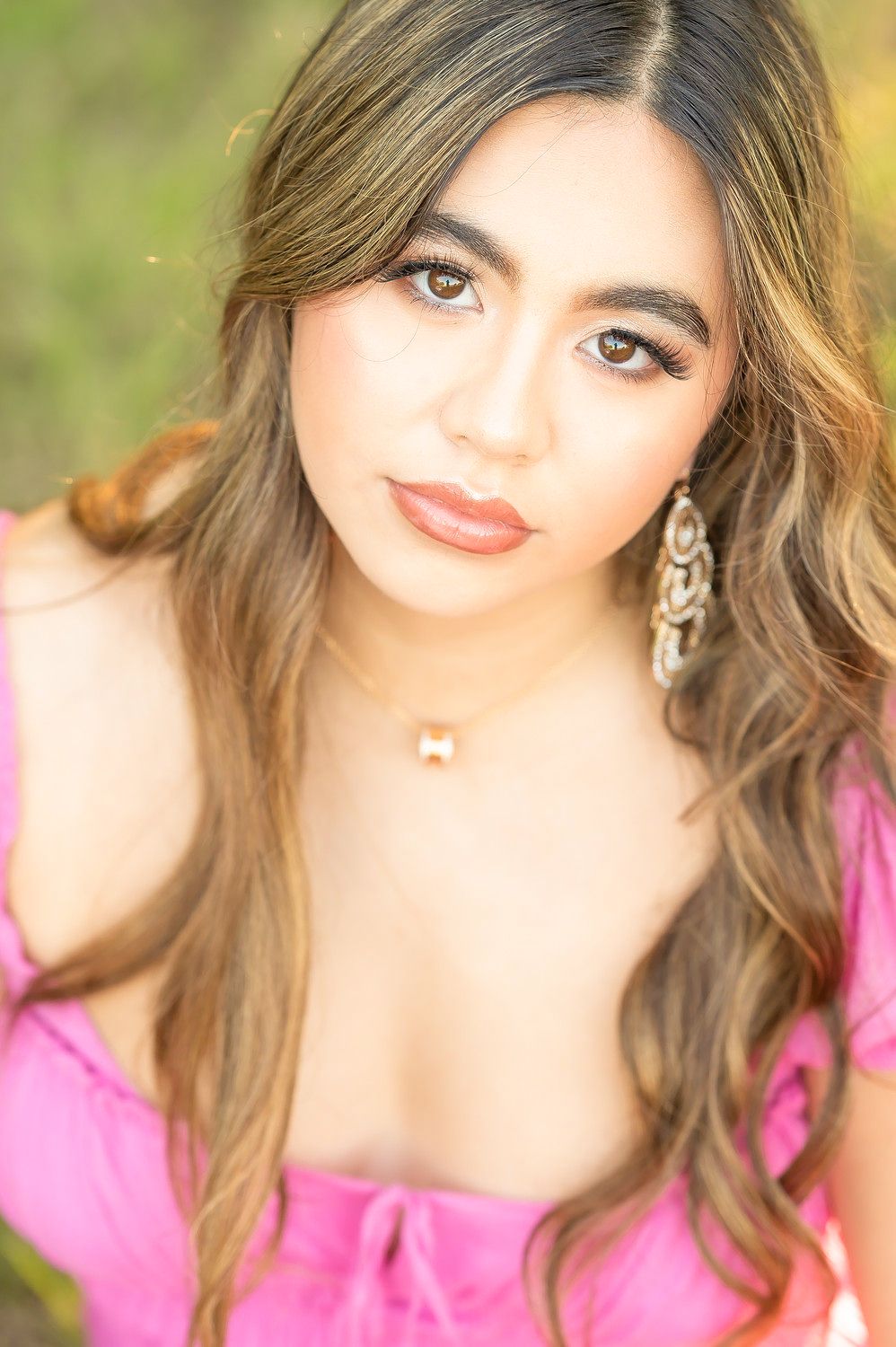 Young woman with long brown hair wearing a pink top, chandelier earrings, and gold necklace poses outdoors.