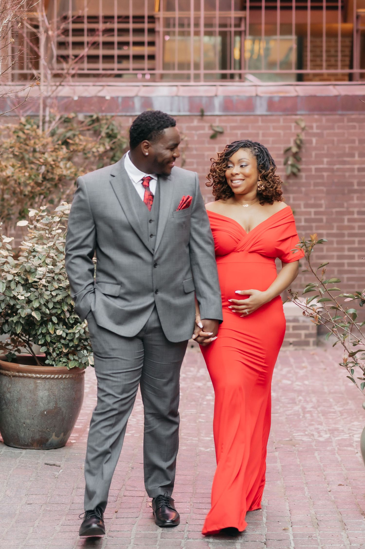 A pregnant woman in a red off-shoulder gown holds hands with a man in a gray suit, sharing a loving glance outdoors.