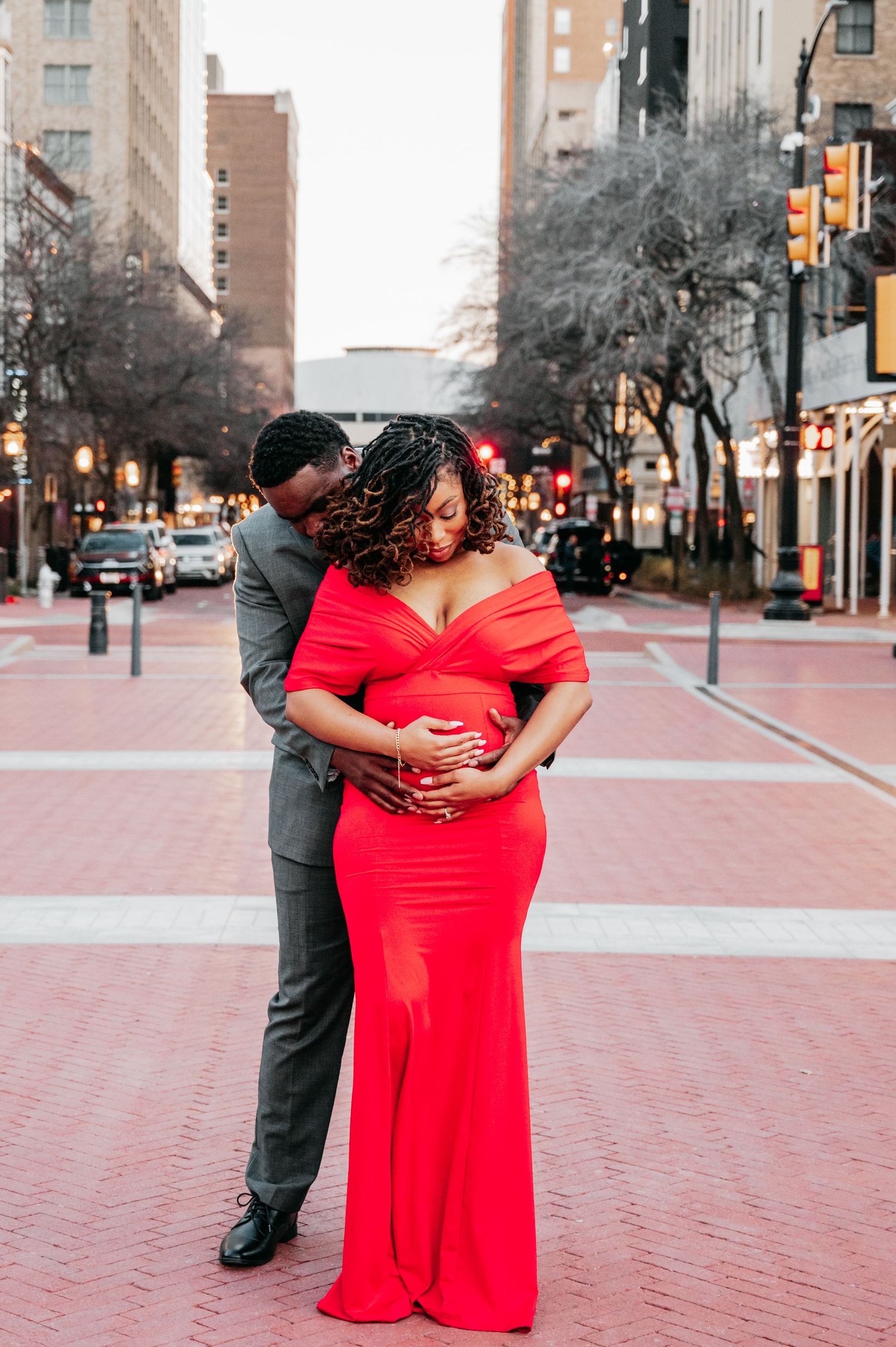 A pregnant woman in a red off-shoulder gown embraced by her partner on a city street during a maternity photoshoot.