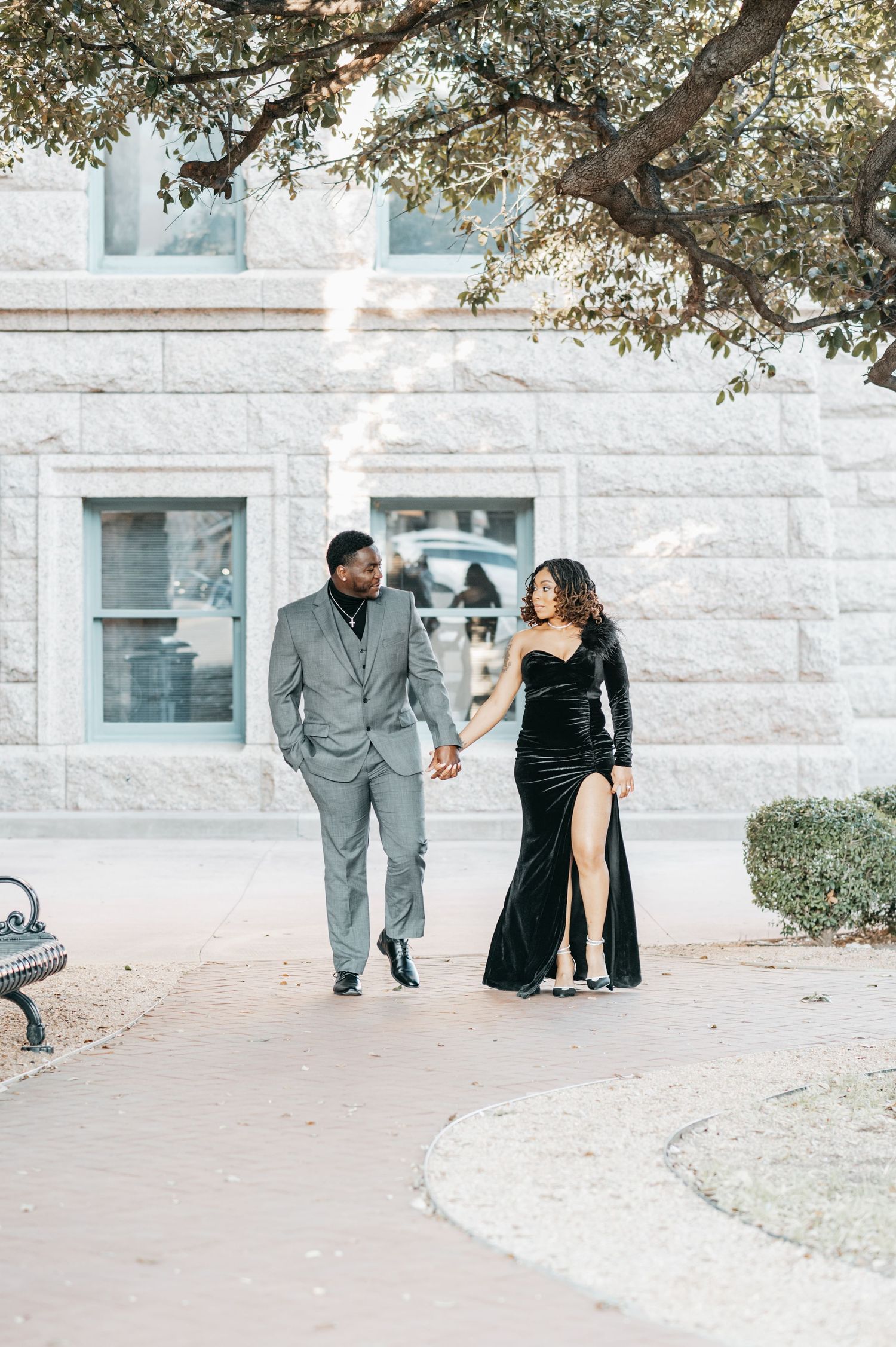 Elegant couple holding hands walking outdoors, man in gray suit and woman in black velvet gown with slit, stone building backdrop.