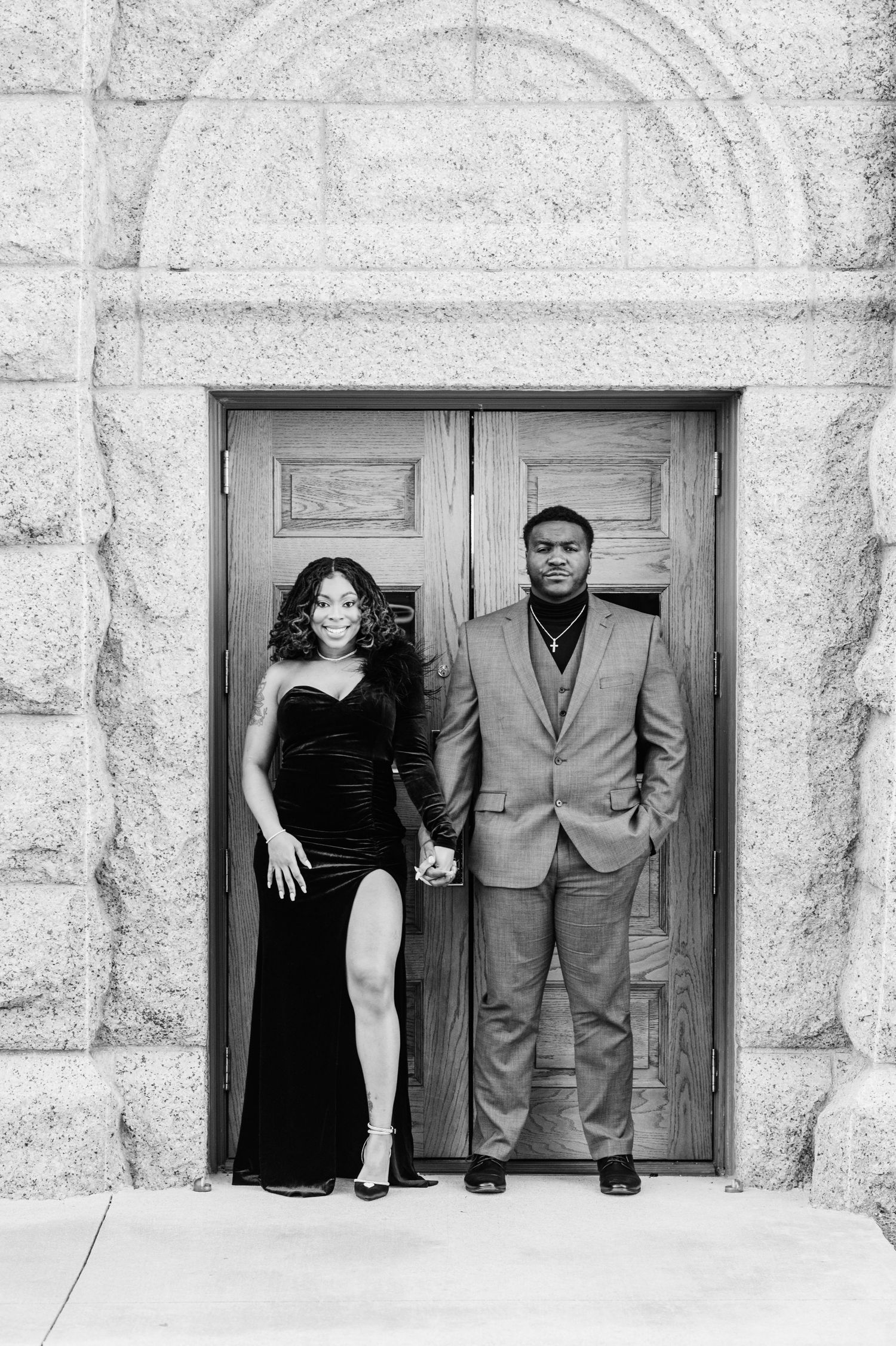 Elegant couple posing in front of ornate wooden doors, woman in black velvet gown, man in suit, black and white photo.
