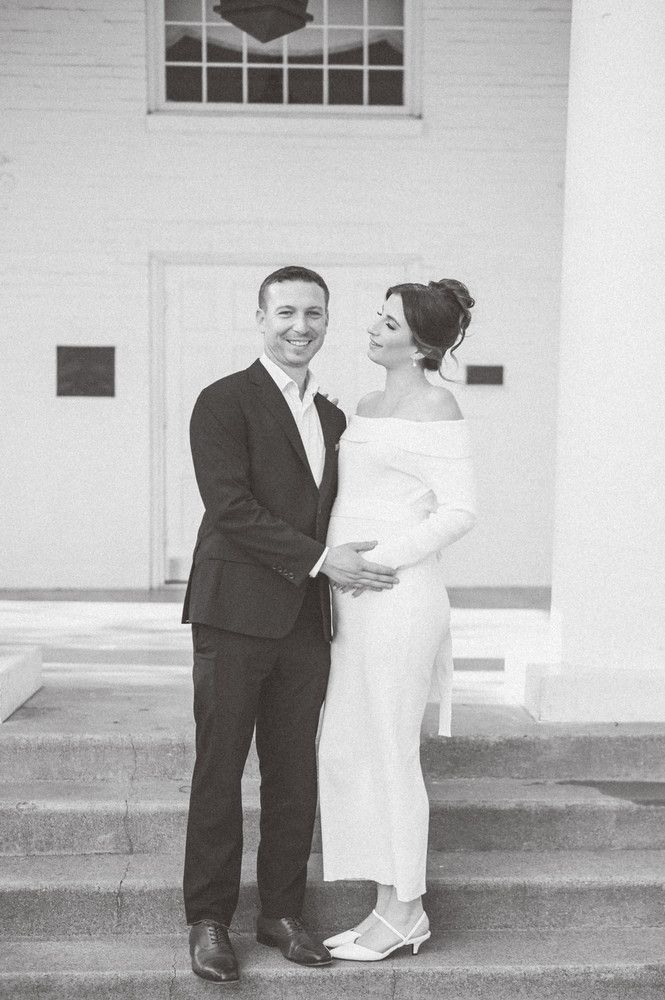 Black and white photo of smiling groom holding pregnant brides belly in white off-shoulder gown on church steps.