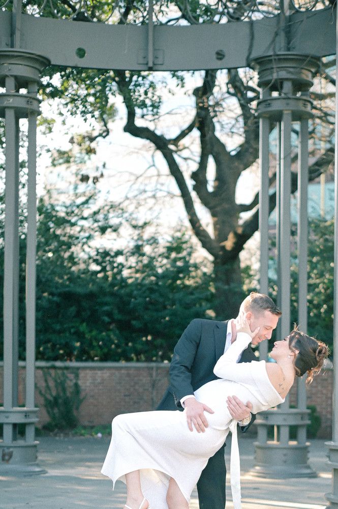 Groom dipping bride in a white off-shoulder dress under a decorative iron gazebo outdoors.