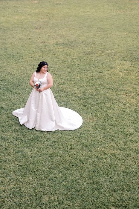 Elegant bride in white satin ball gown with long train stands on green grass holding bouquet.
