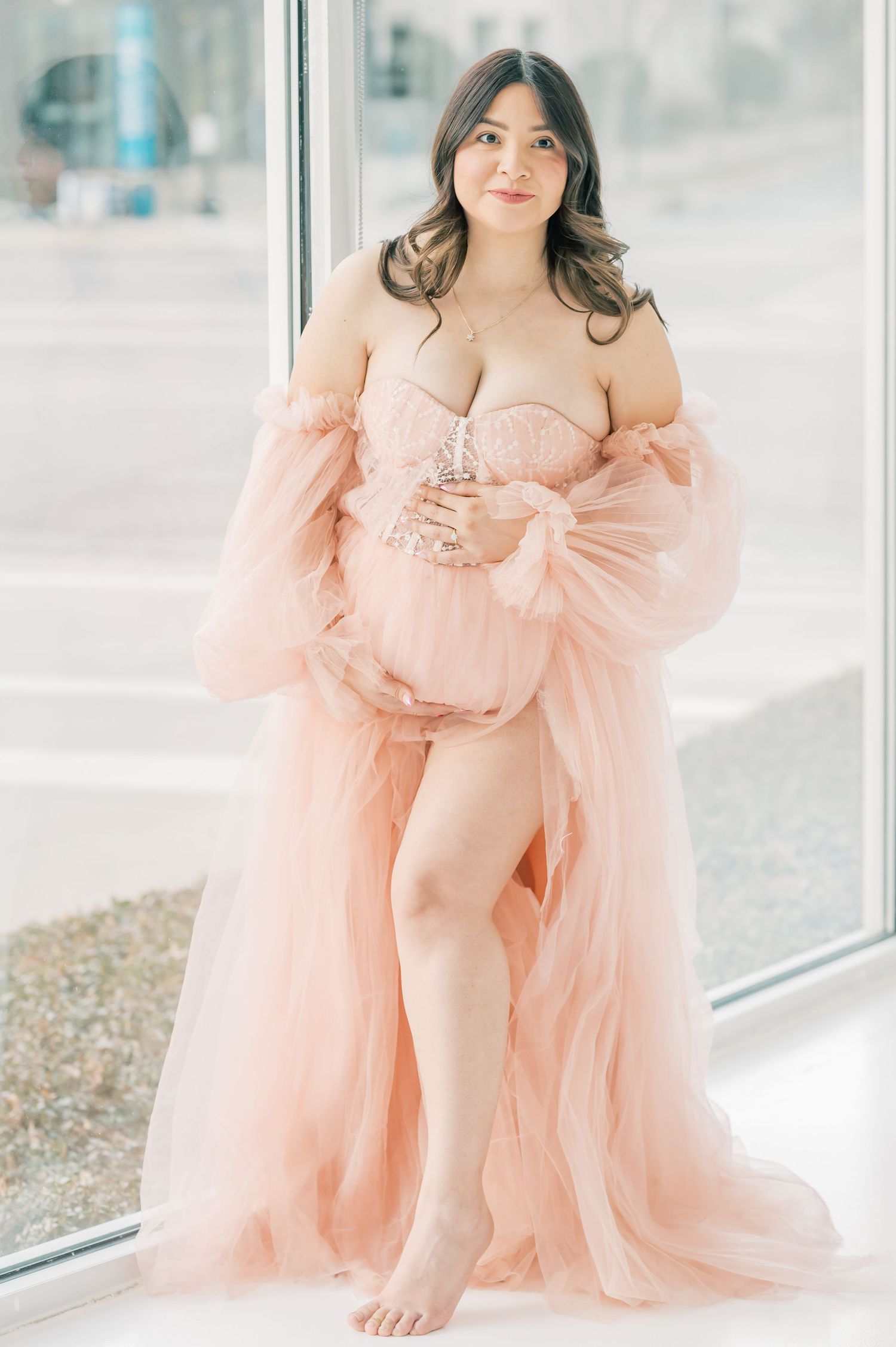 Pregnant woman in blush pink tulle maternity gown posing near large window for elegant maternity photo session.