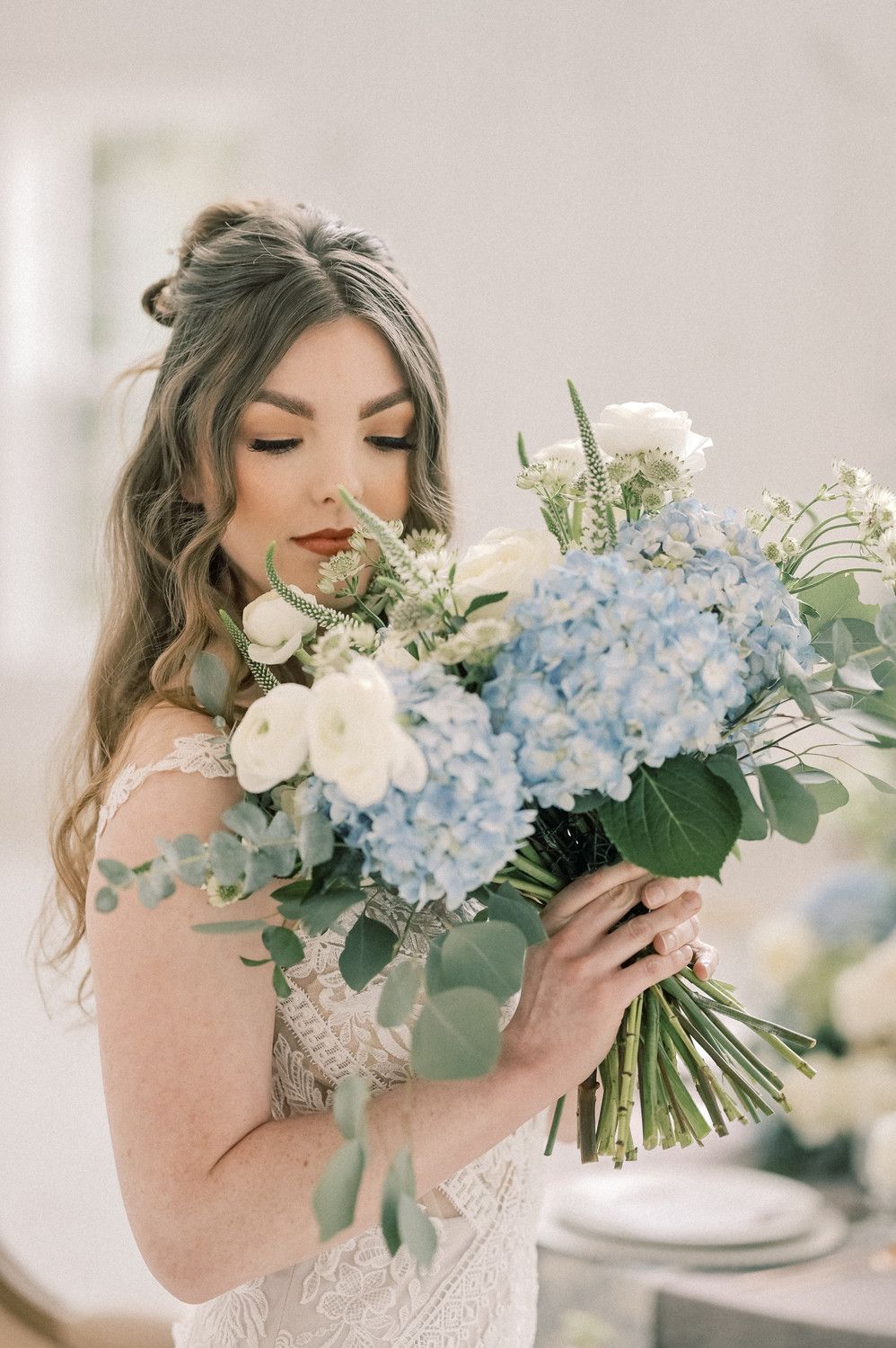 Bride in lace gown smelling a bouquet of blue hydrangeas and white roses with eucalyptus greenery.