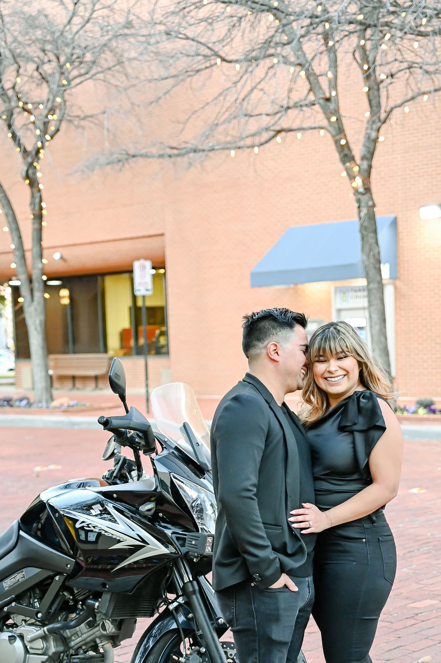 Couple in black outfits laughing together beside a motorcycle on a brick street with bare winter trees.