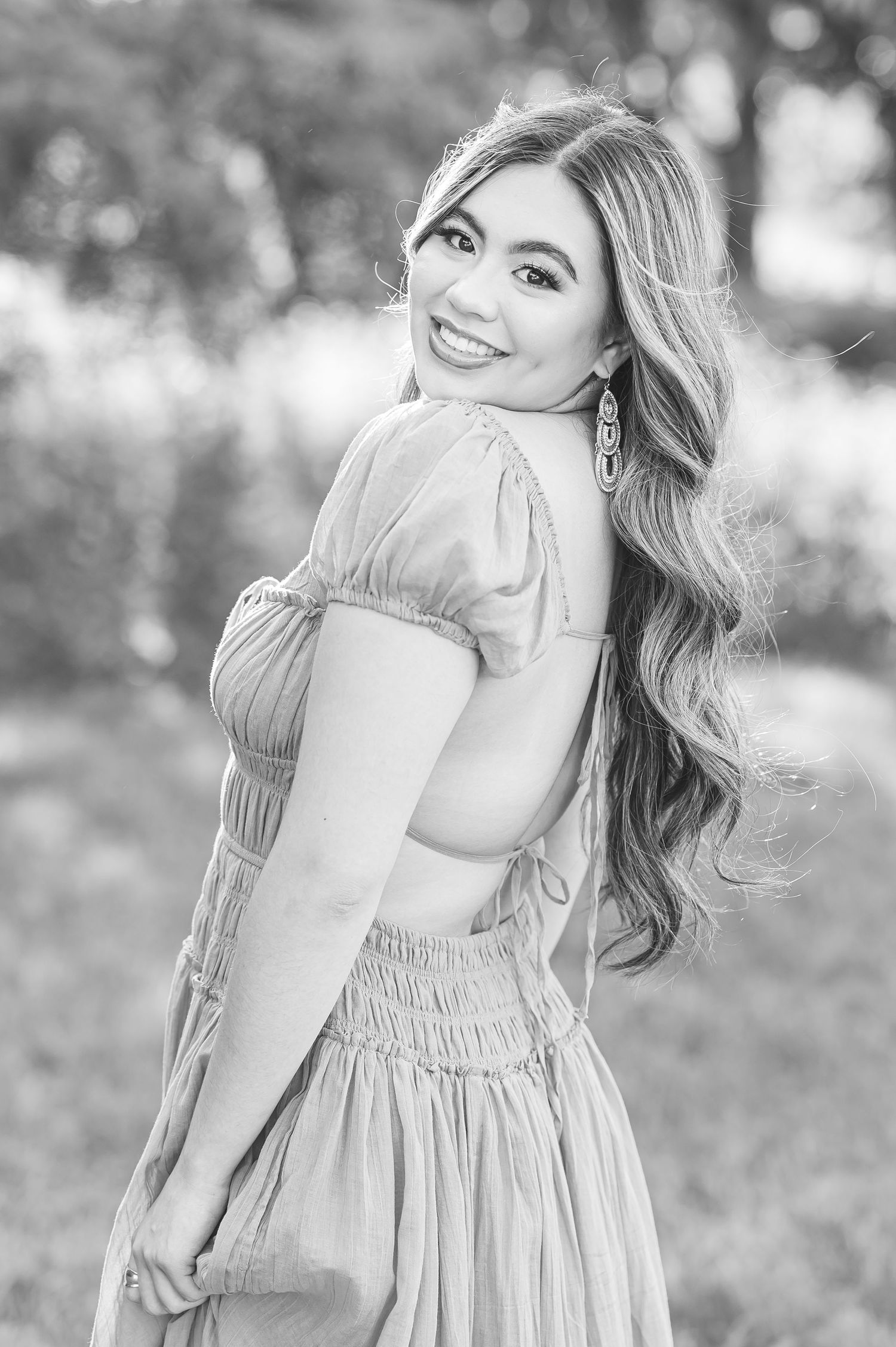 Black and white portrait of a smiling woman in a vintage-style dress with long wavy hair posing outdoors.