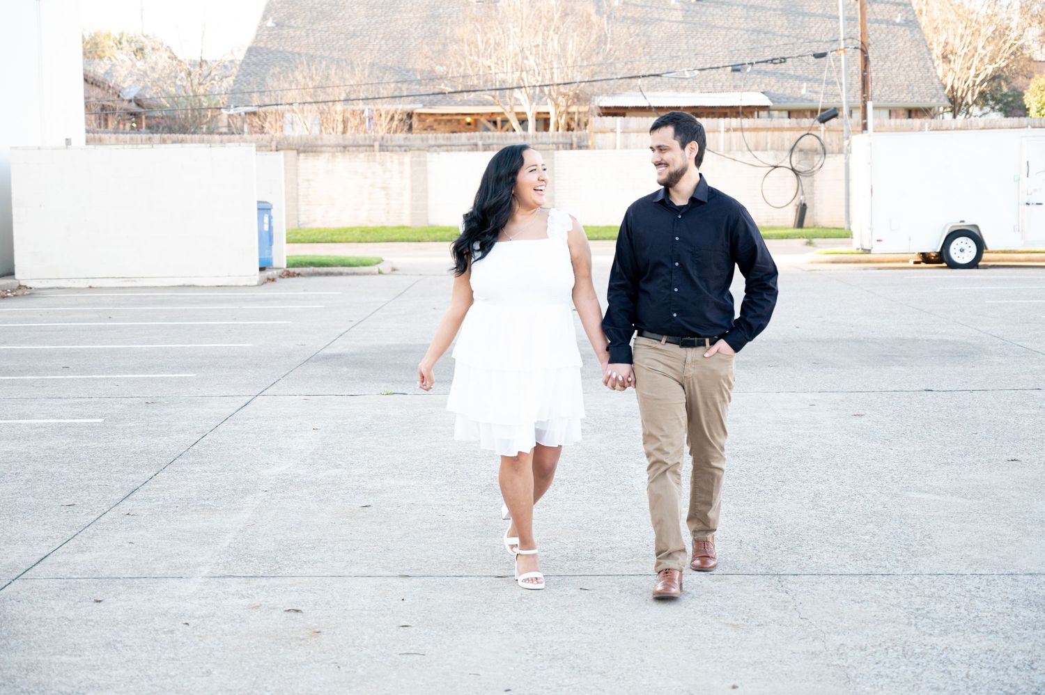 A couple walks together in an outdoor parking area, with the person in white wearing a dress and the other in dark attire.