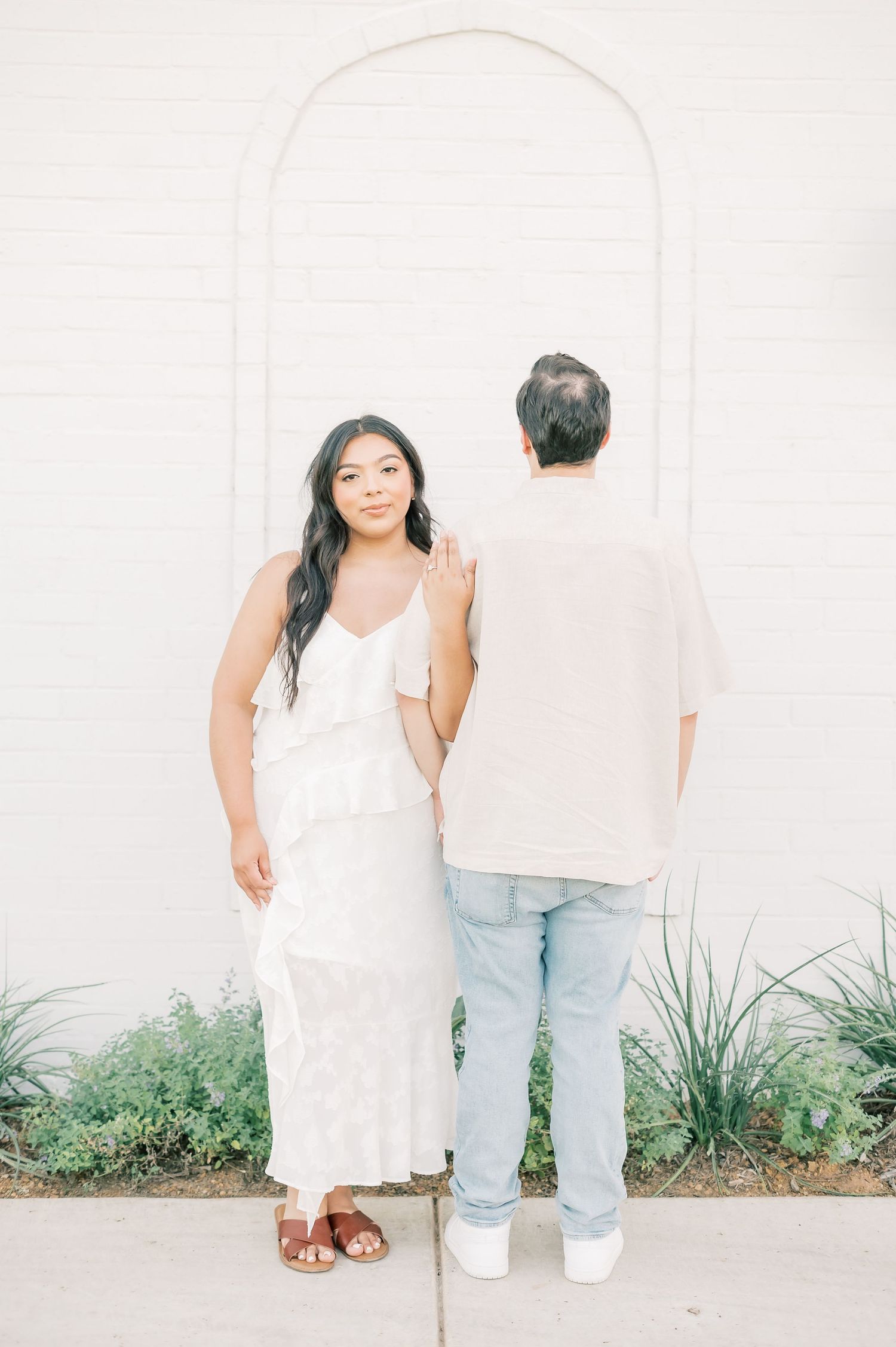 A romantic couple in white outfits poses against a minimalist white wall with green plants at the base.