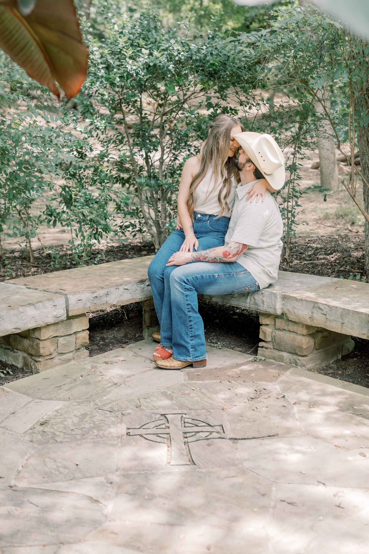 Couple sitting close on a stone bench surrounded by lush greenery, with a cross etched in the ground below.