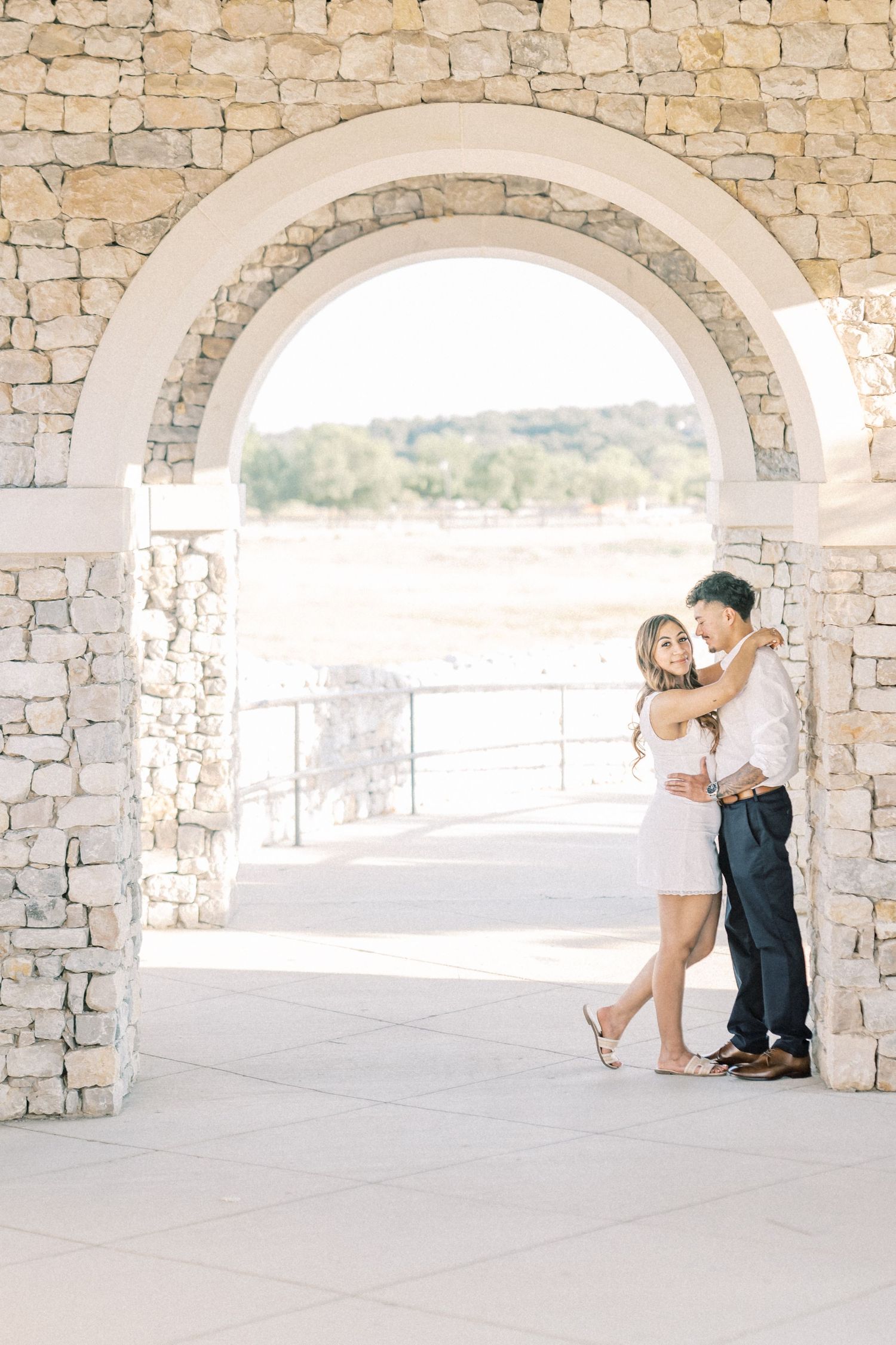 Couple embracing under a stone arch during their engagement session at an outdoor venue.