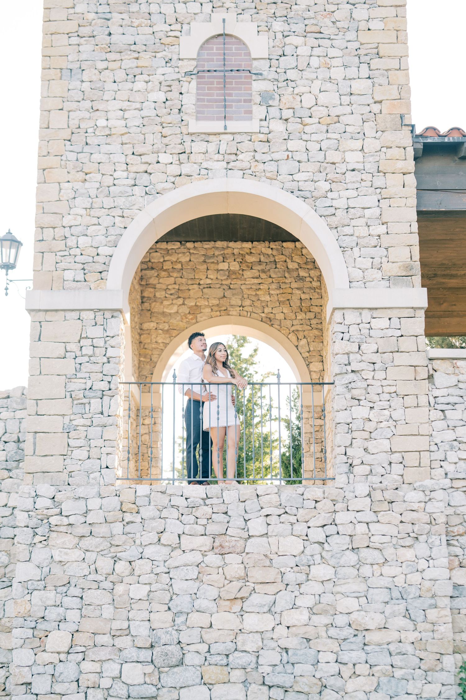 An engaged couple stands together under a stone archway of a rustic building, gazing into the distance.