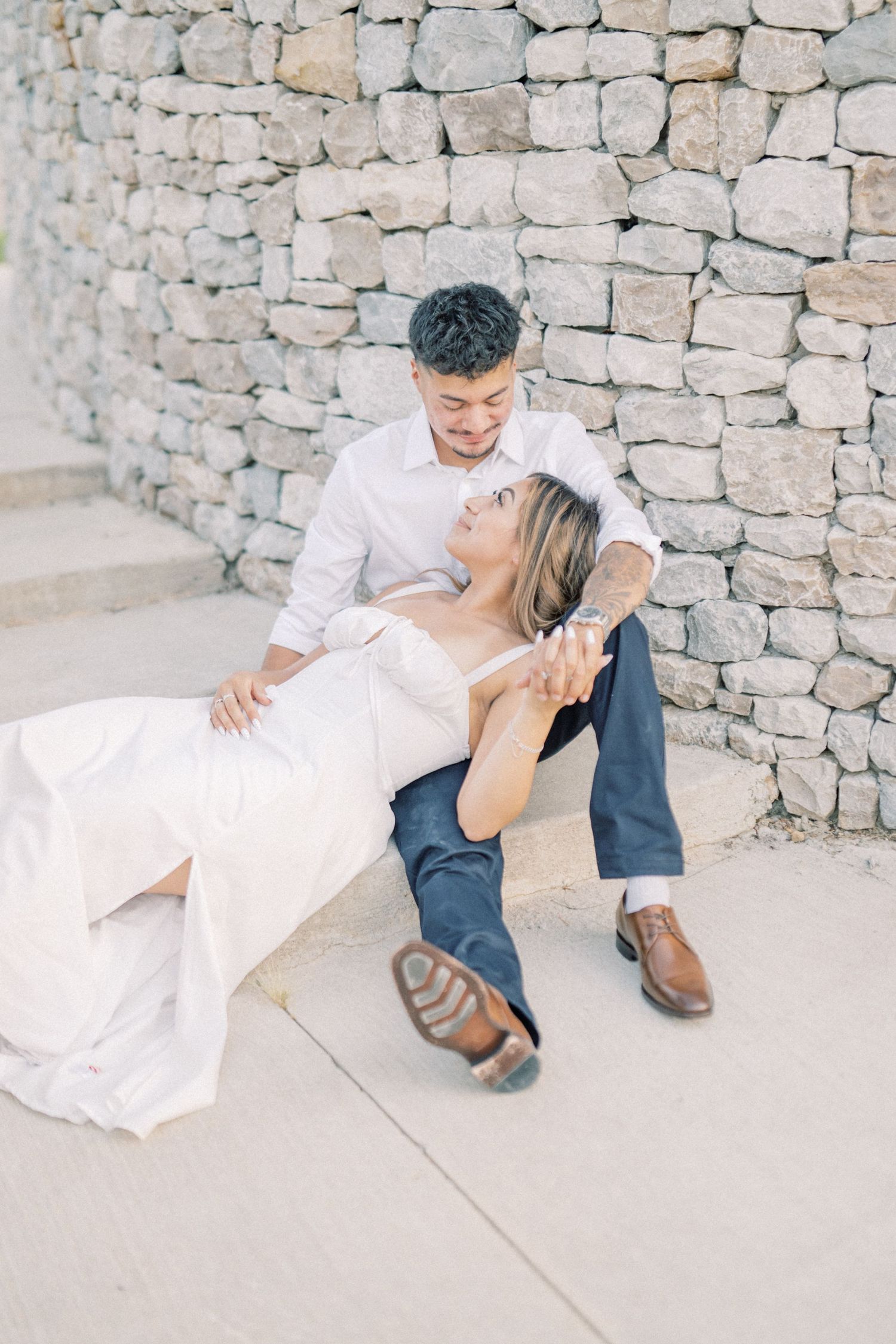 A couple shares a romantic, intimate moment seated against a textured stone wall during their engagement session.