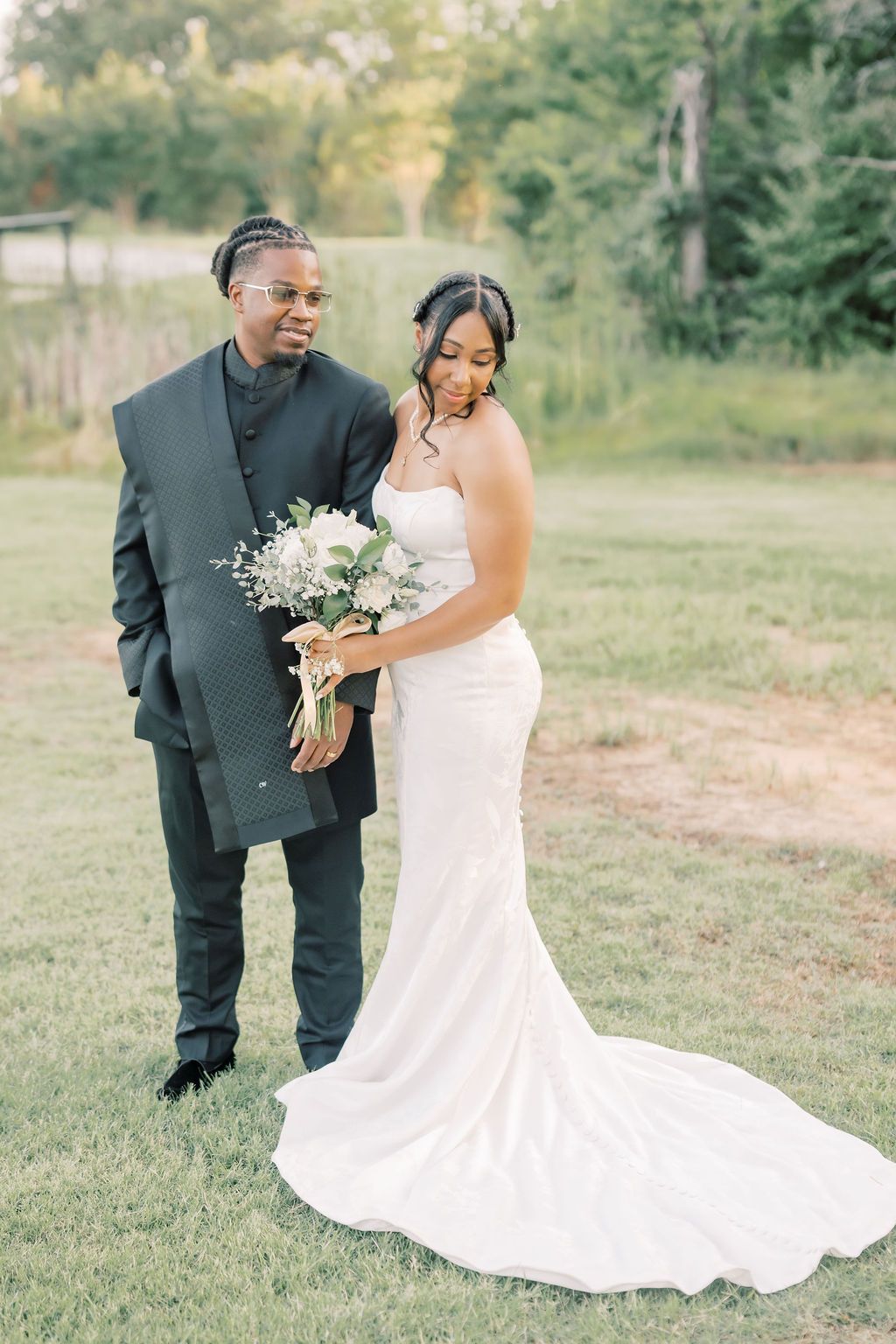 A bride in a white gown holds a floral bouquet beside a groom in a black suit, posing outdoors on green grass.