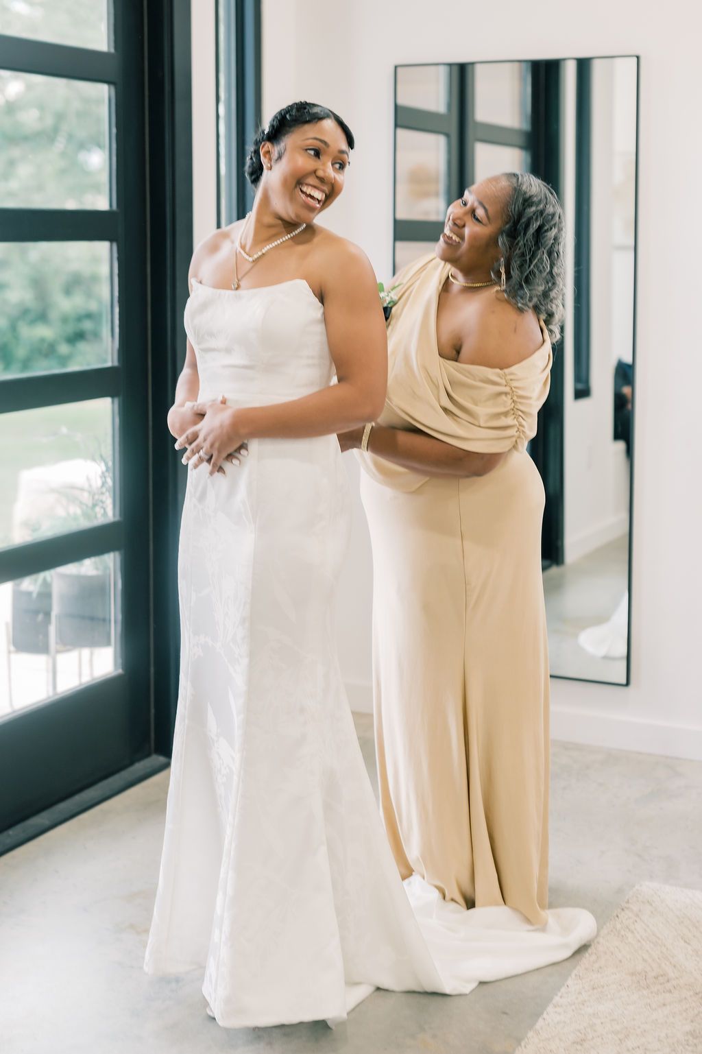 Bride in white strapless gown smiles as older woman in gold dress helps her prepare near elegant black French doors.
