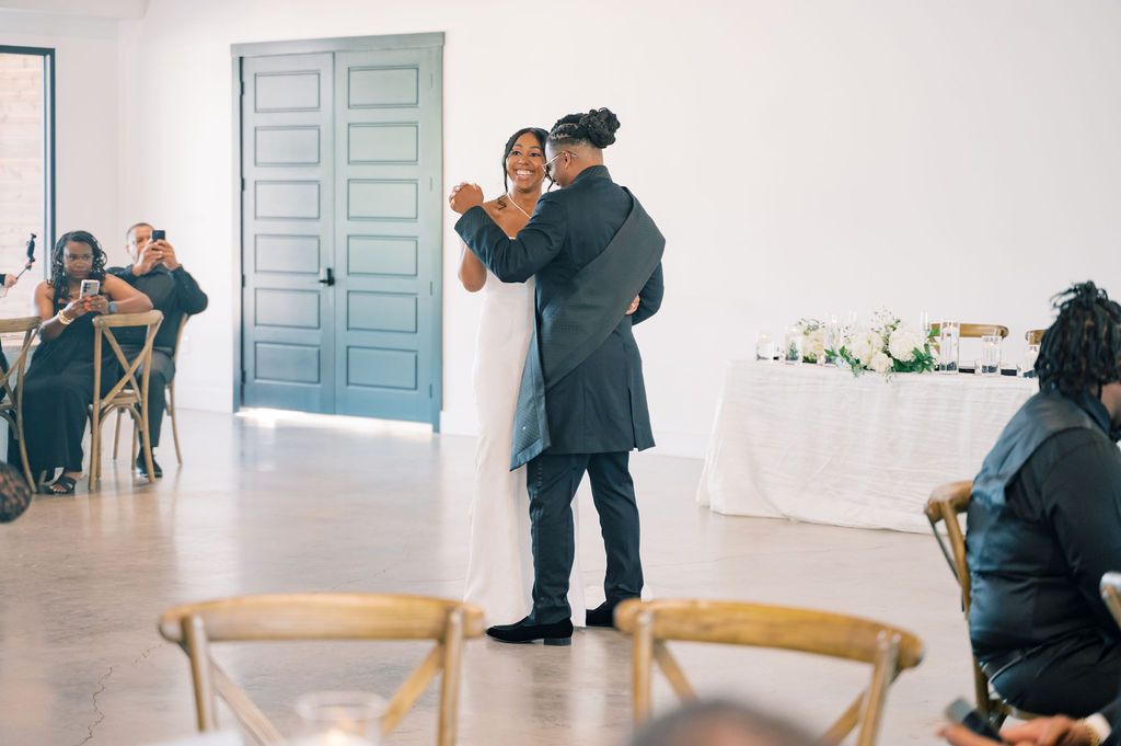Newlyweds share their first dance in a modern white venue with teal doors and seated wedding guests.