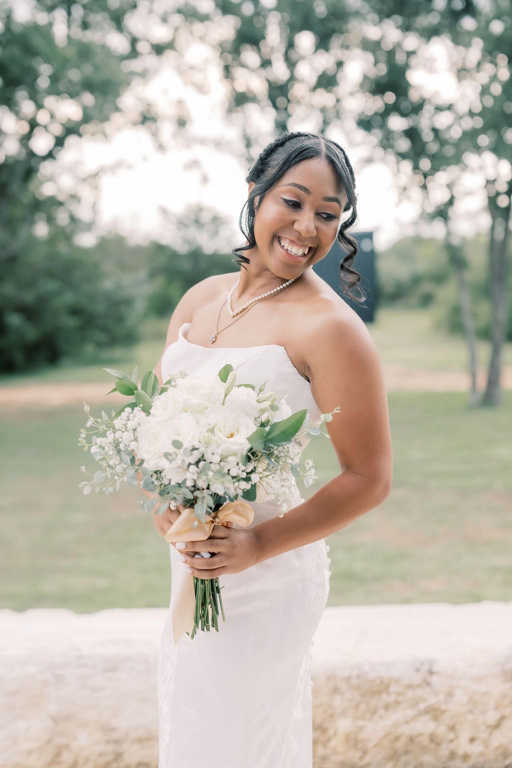 Smiling bride in strapless white gown holding elegant white floral bouquet outdoors on her wedding day.