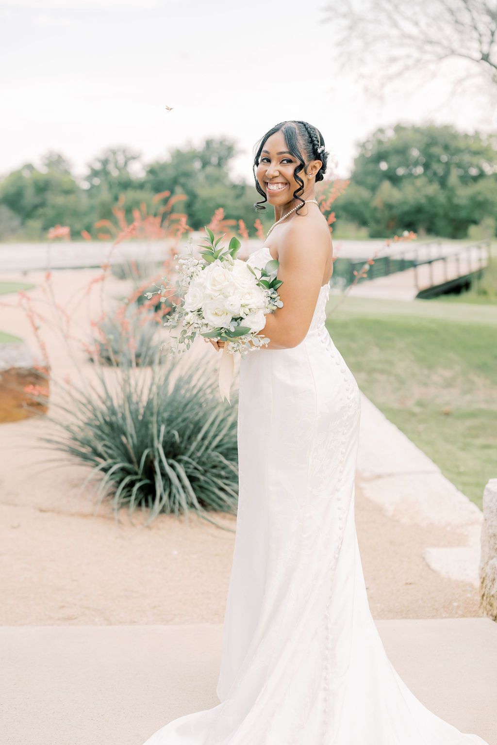 Smiling bride in a white fitted gown holds a white floral bouquet outdoors near lush greenery and a scenic pathway.