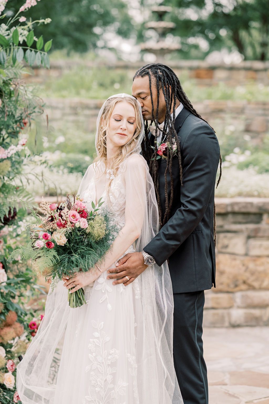 Interracial bride and groom sharing a tender moment with a colorful bouquet at an outdoor garden wedding.