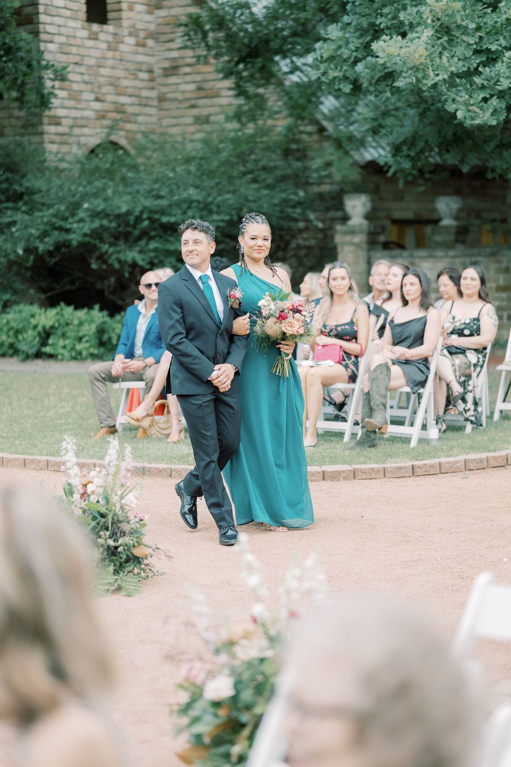 Groomsman escorts bridesmaid in teal gown down outdoor wedding aisle, guests seated in white chairs.