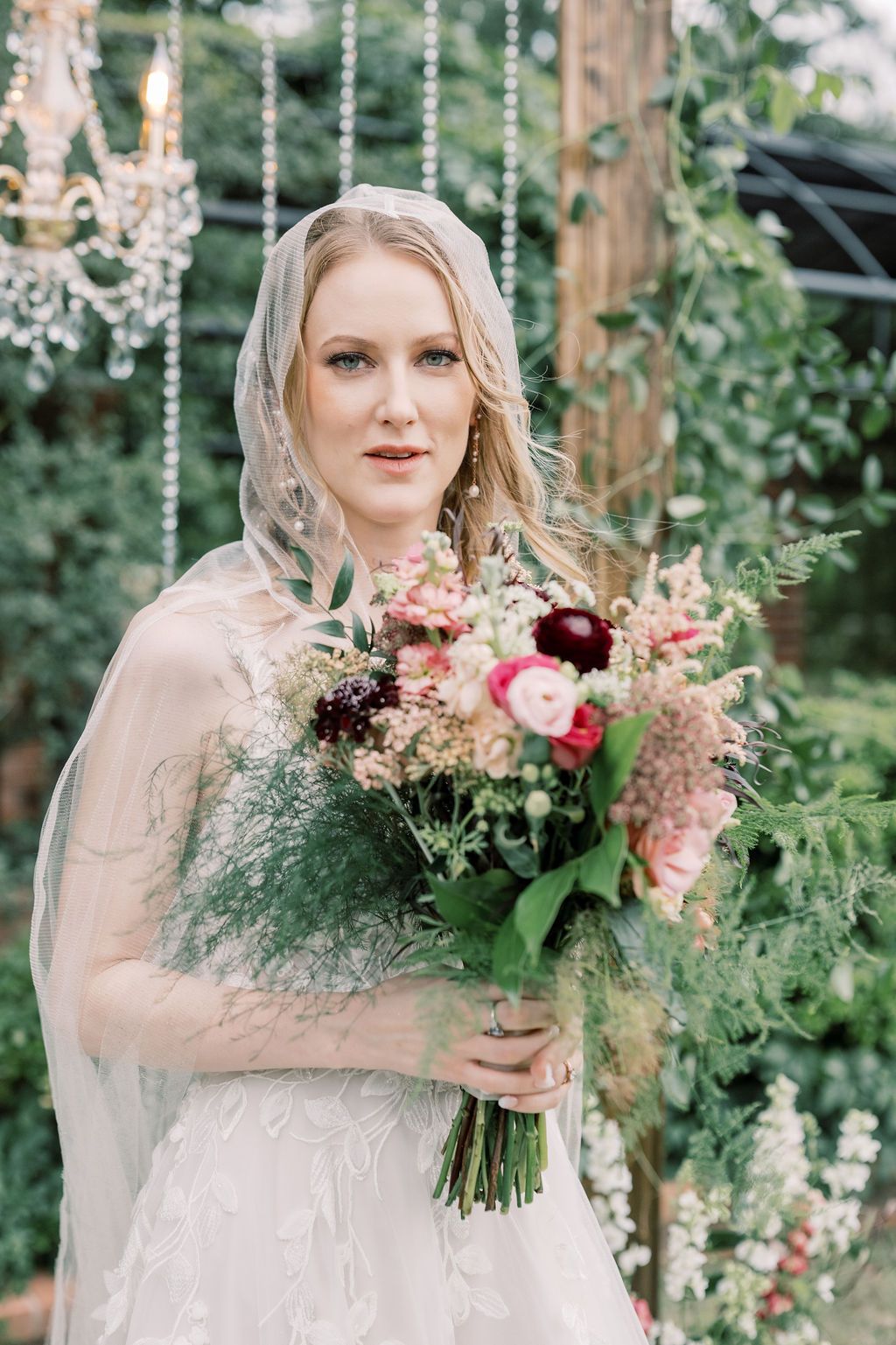 Elegant bride in lace gown and veil holding lush floral bouquet with pink and burgundy blooms in garden setting.
