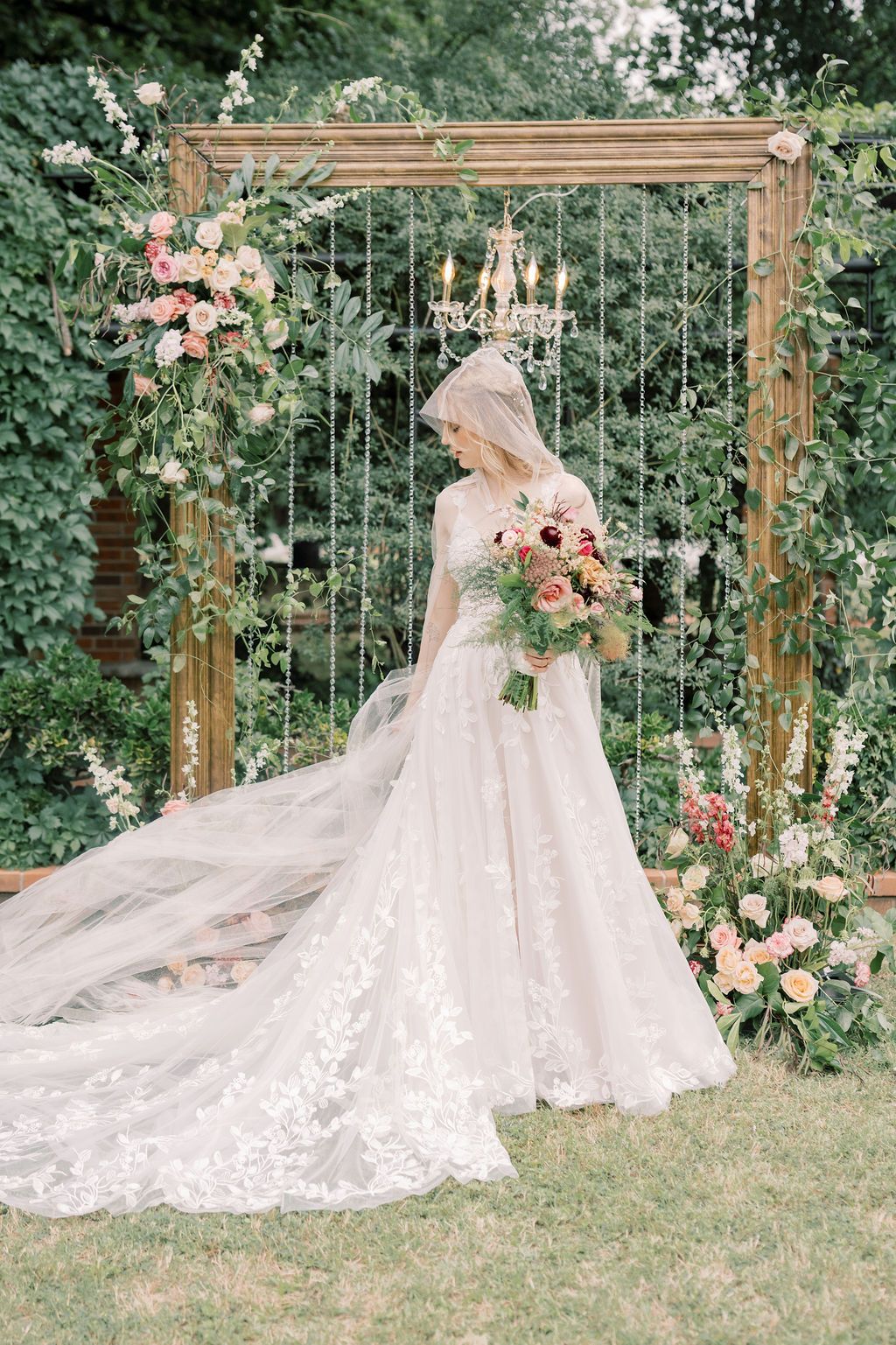 Bride in lace ballgown holding bouquet stands before floral wooden arch with chandelier in lush garden setting.
