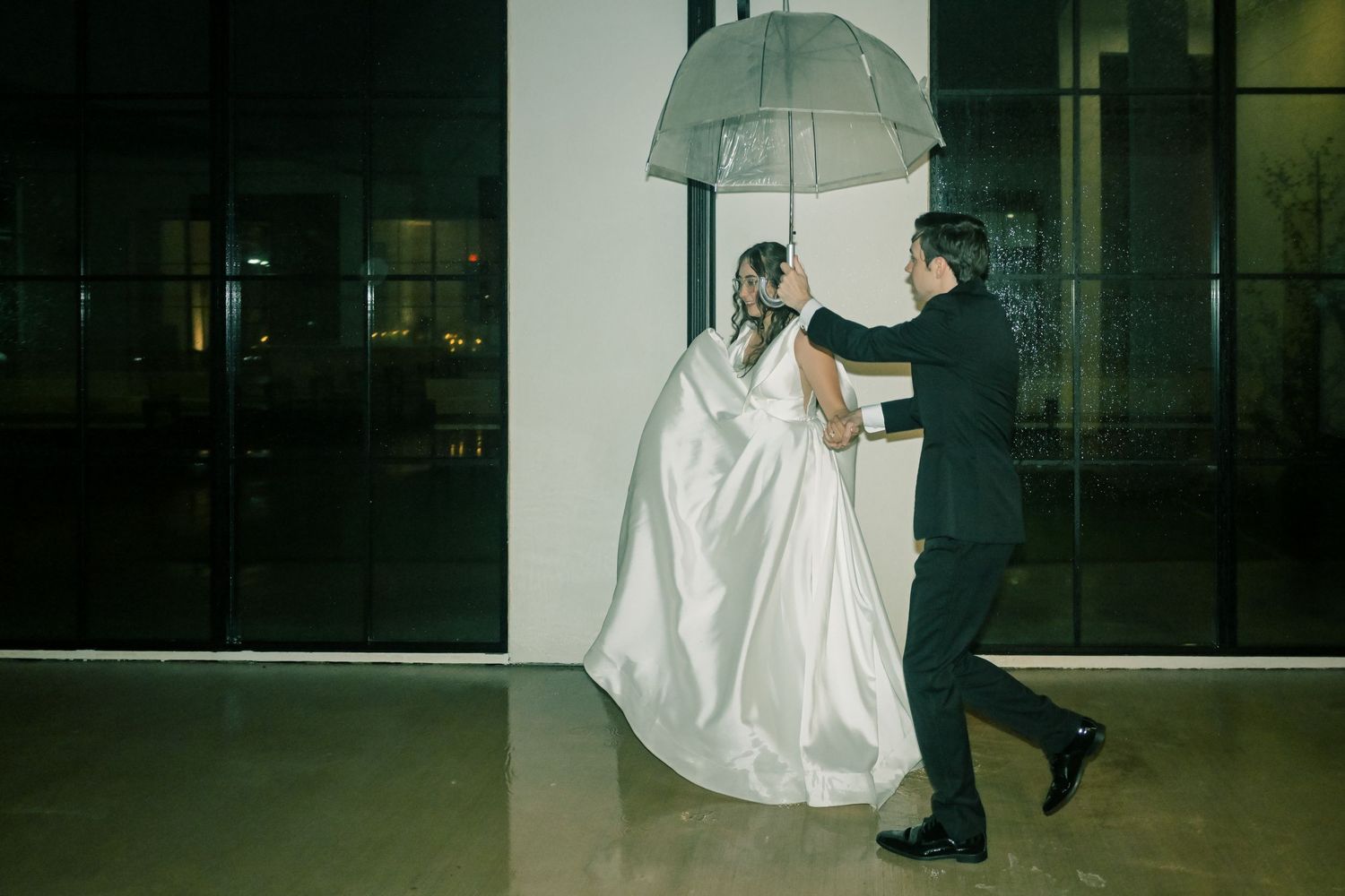 Bride and groom walk together under a clear umbrella outside a modern venue on a rainy wedding night.