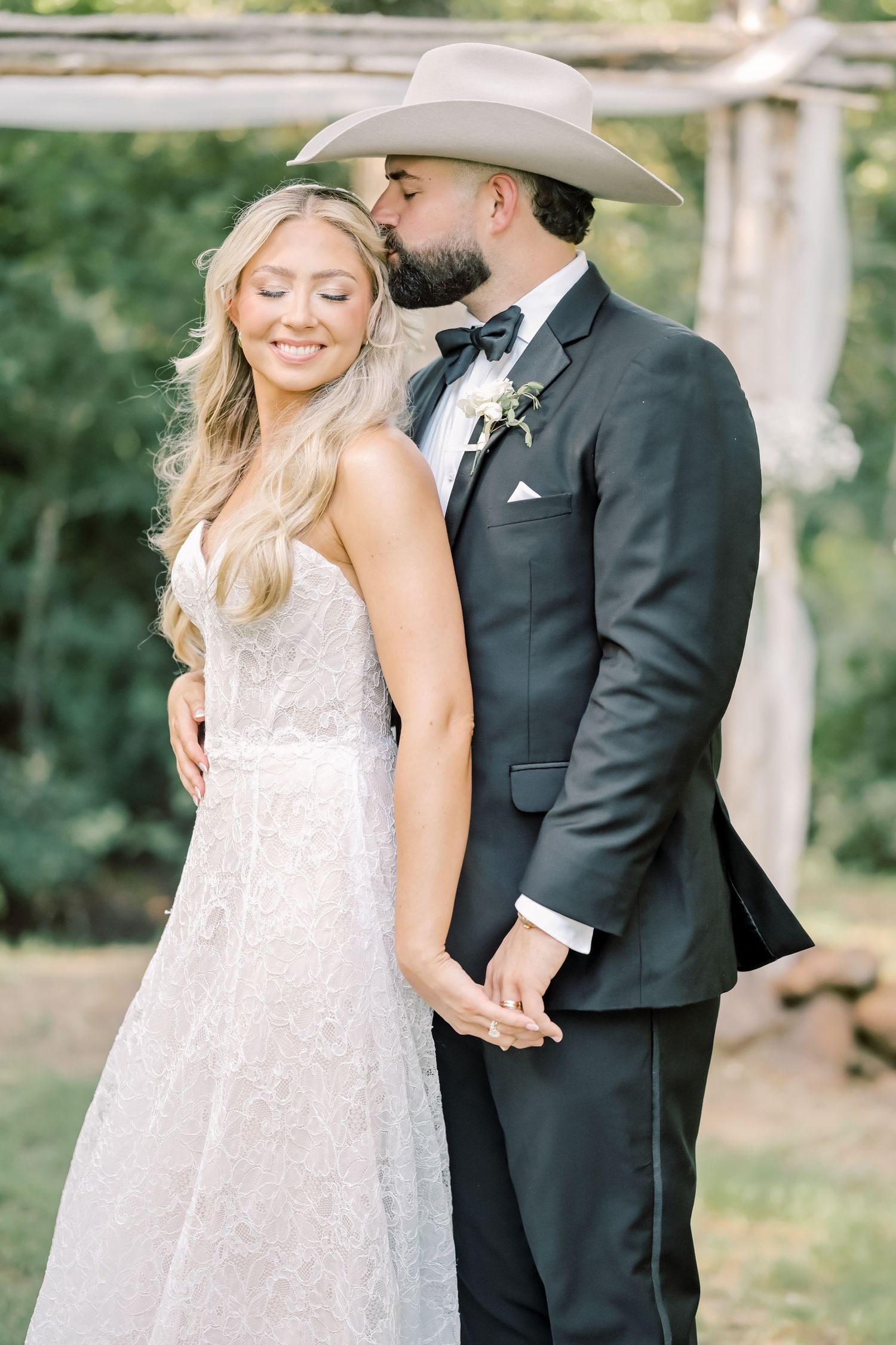 Groom in black tuxedo and cowboy hat kissing smiling bride in lace wedding dress outdoors.
