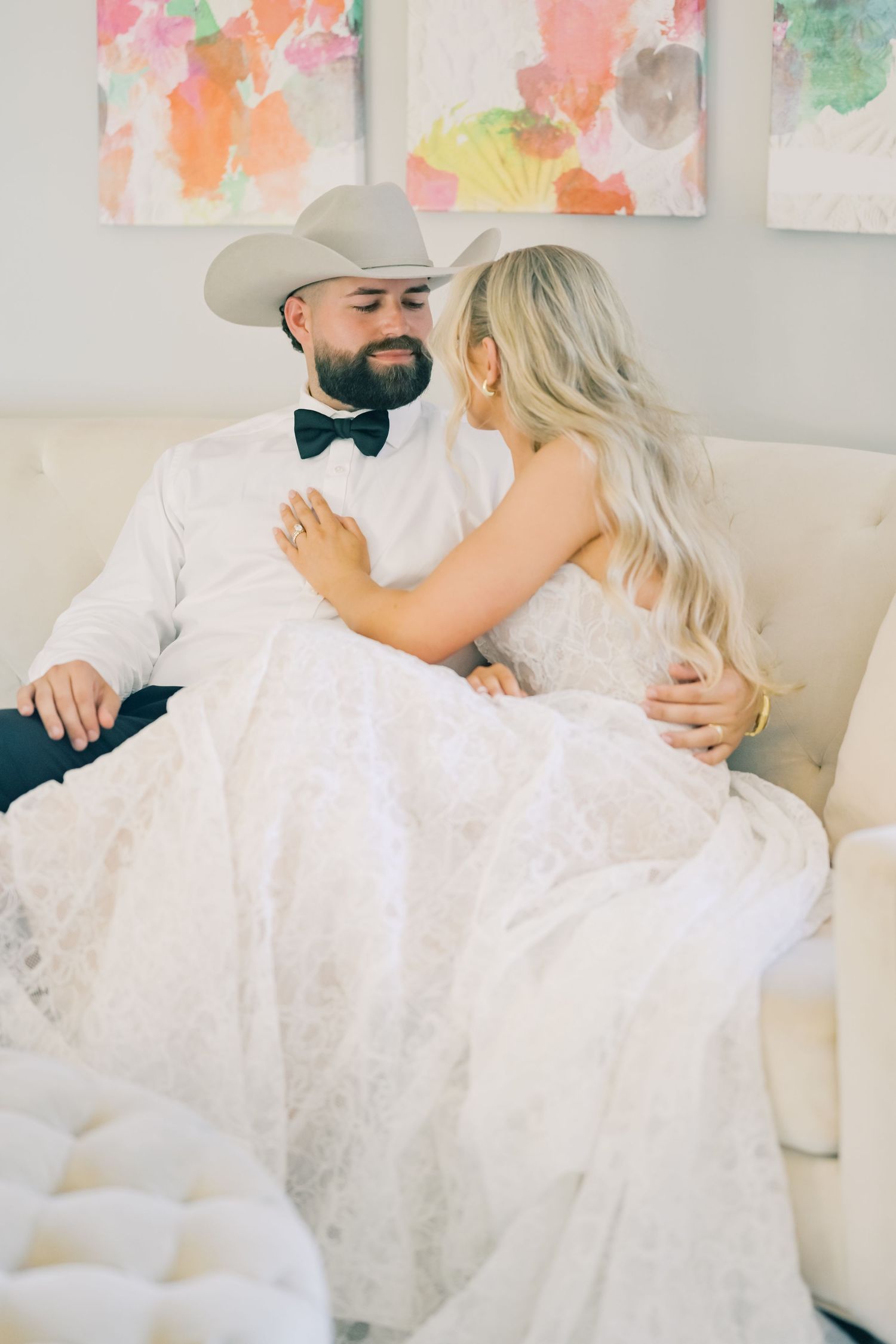 A bride in a lace gown sits with her bearded groom in a cowboy hat on a white sofa.