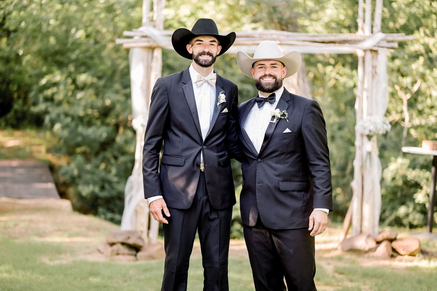 Two groomsmen wearing black suits and cowboy hats smile together outdoors near rustic wooden wedding arch.