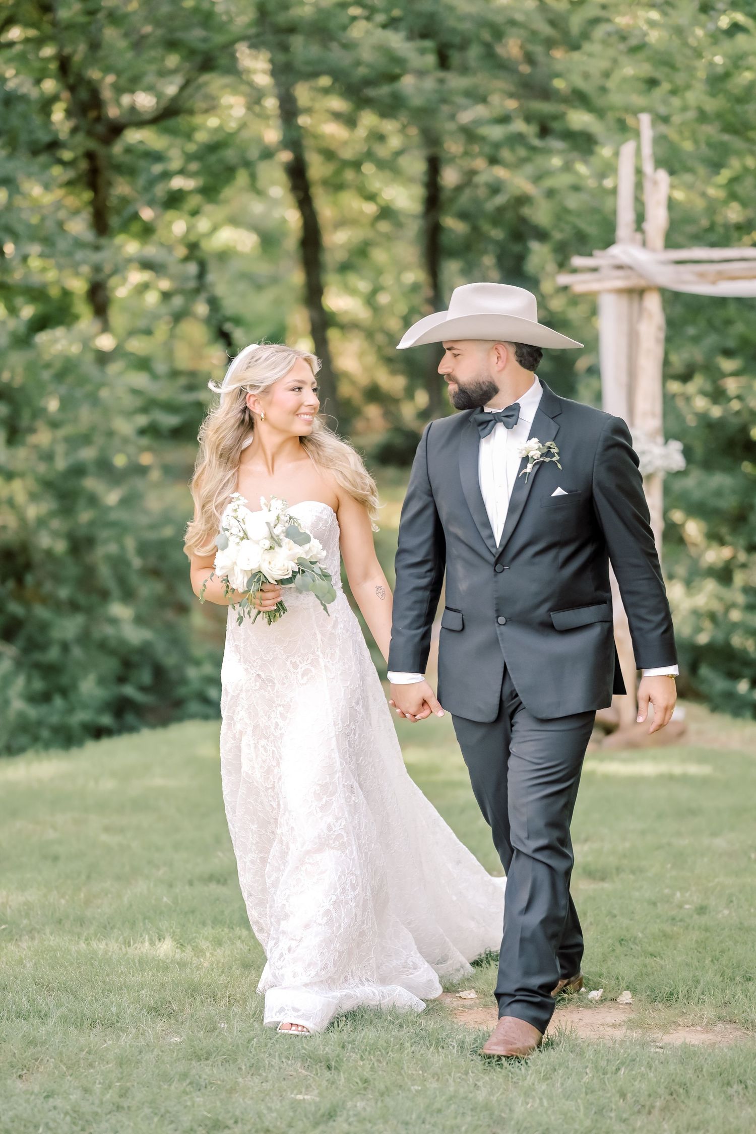 Bride and groom walk hand-in-hand outdoors, she holds white bouquet, he wears cowboy hat.