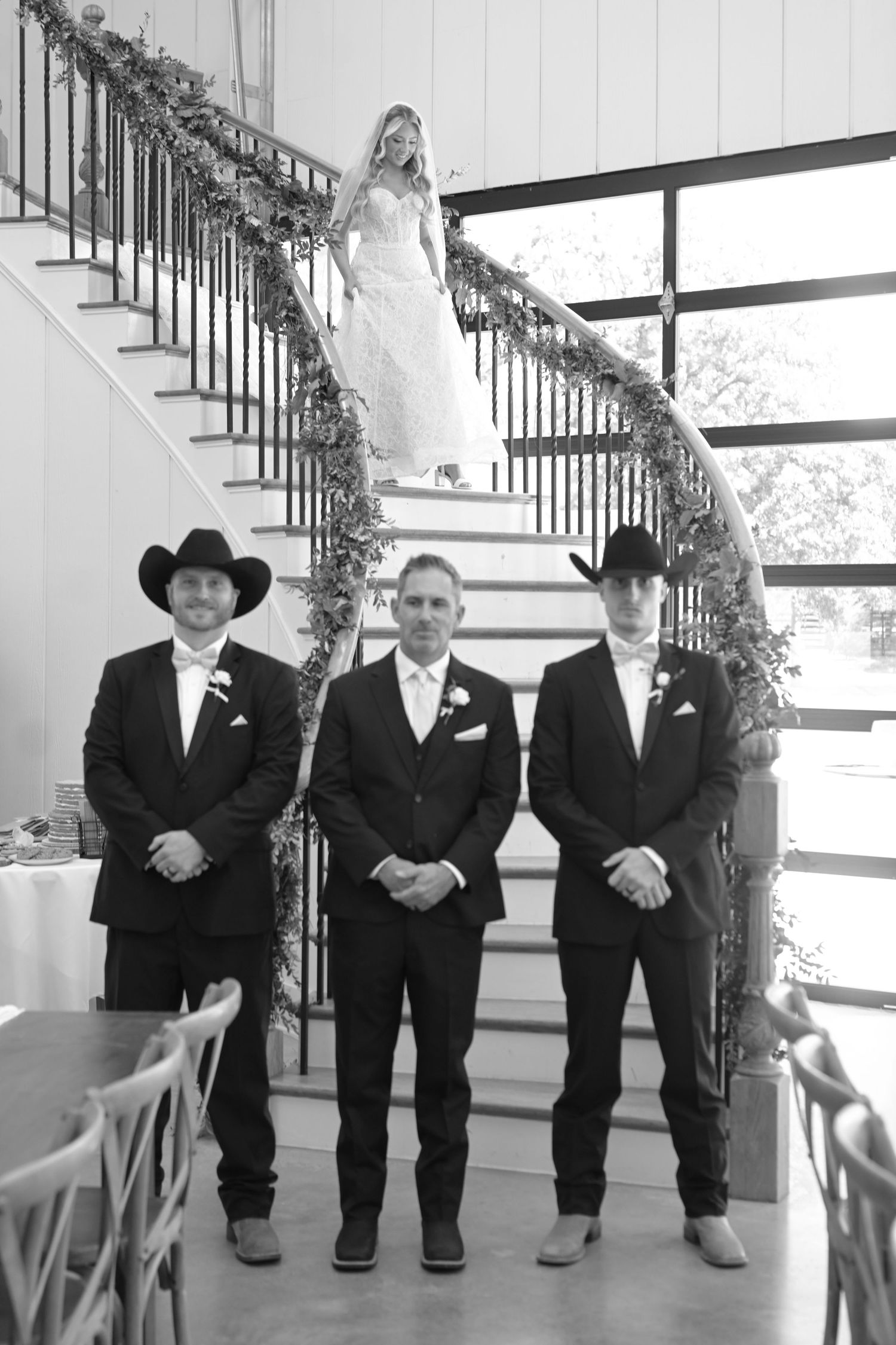 Bride descends floral staircase behind three groomsmen in black suits and cowboy hats at rustic wedding venue.